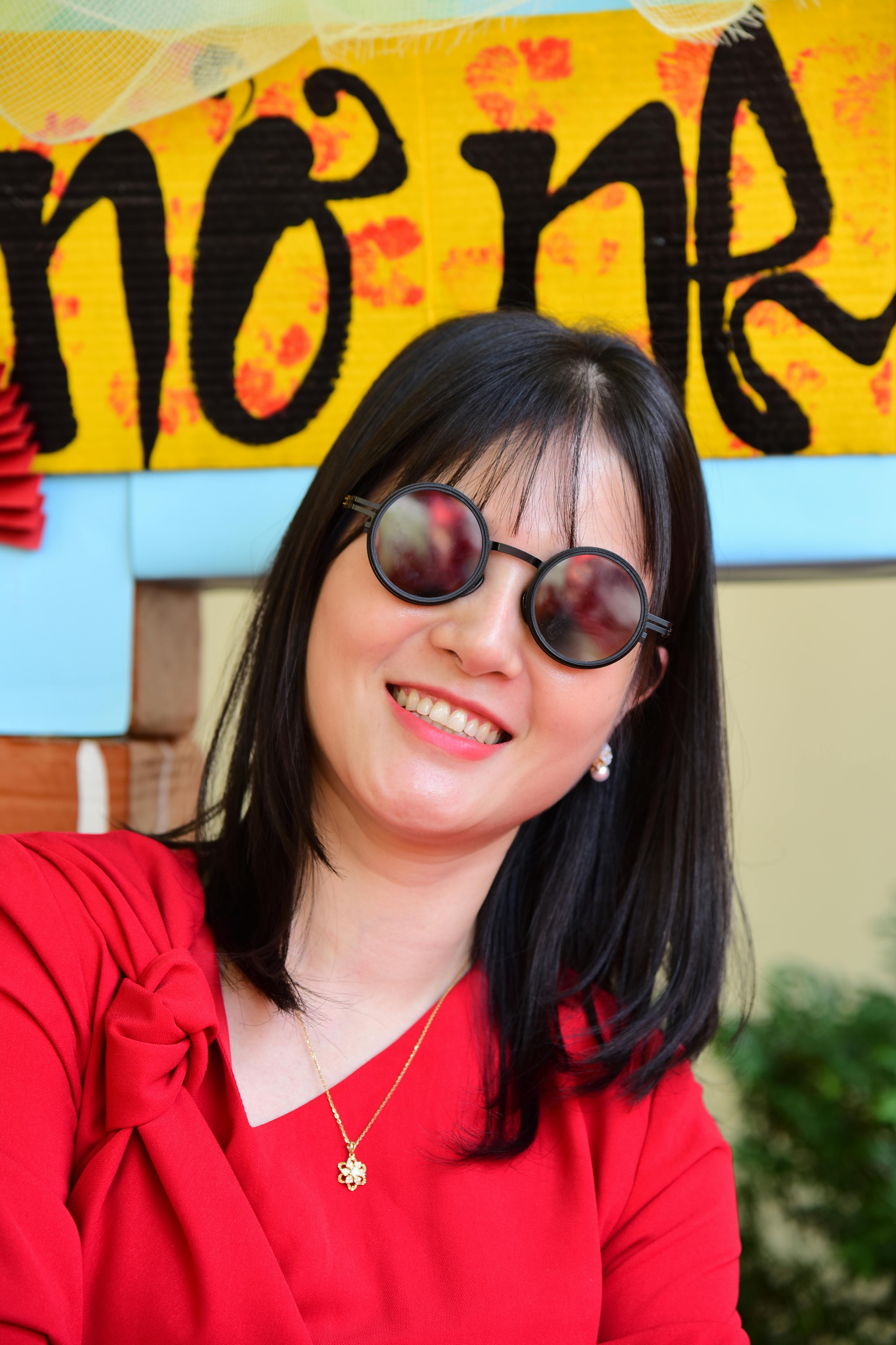 A cheerful woman in a red dress wearing round sunglasses, smiling warmly.