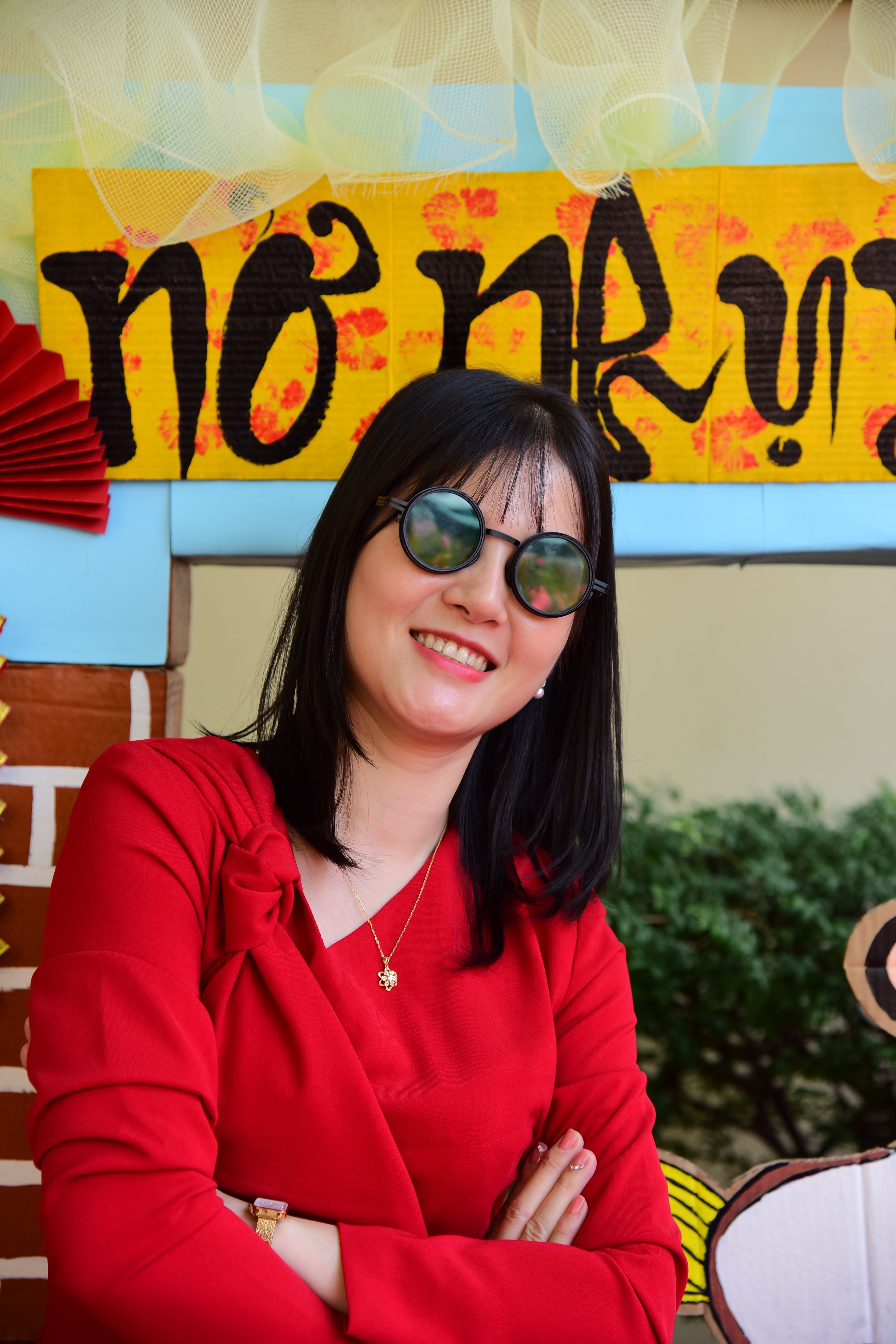 A stylish woman in red wearing sunglasses stands by a colorful event sign.