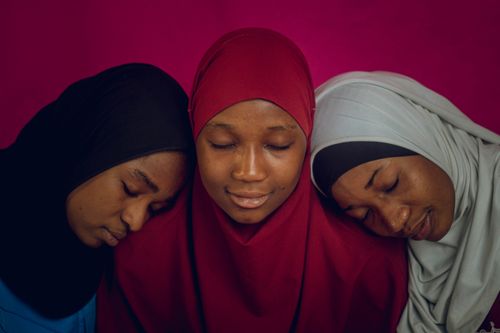Free A serene portrait of three women in hijabs resting with eyes closed against a vibrant pink background. Stock Photo