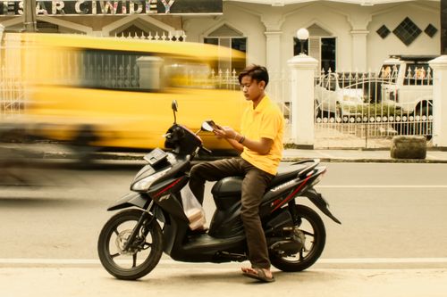 Free A young man reads on his motorbike as a yellow van speeds by, illustrating urban life. Stock Photo