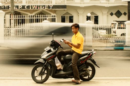 A young man sits on a scooter reading while parked by a busy street.