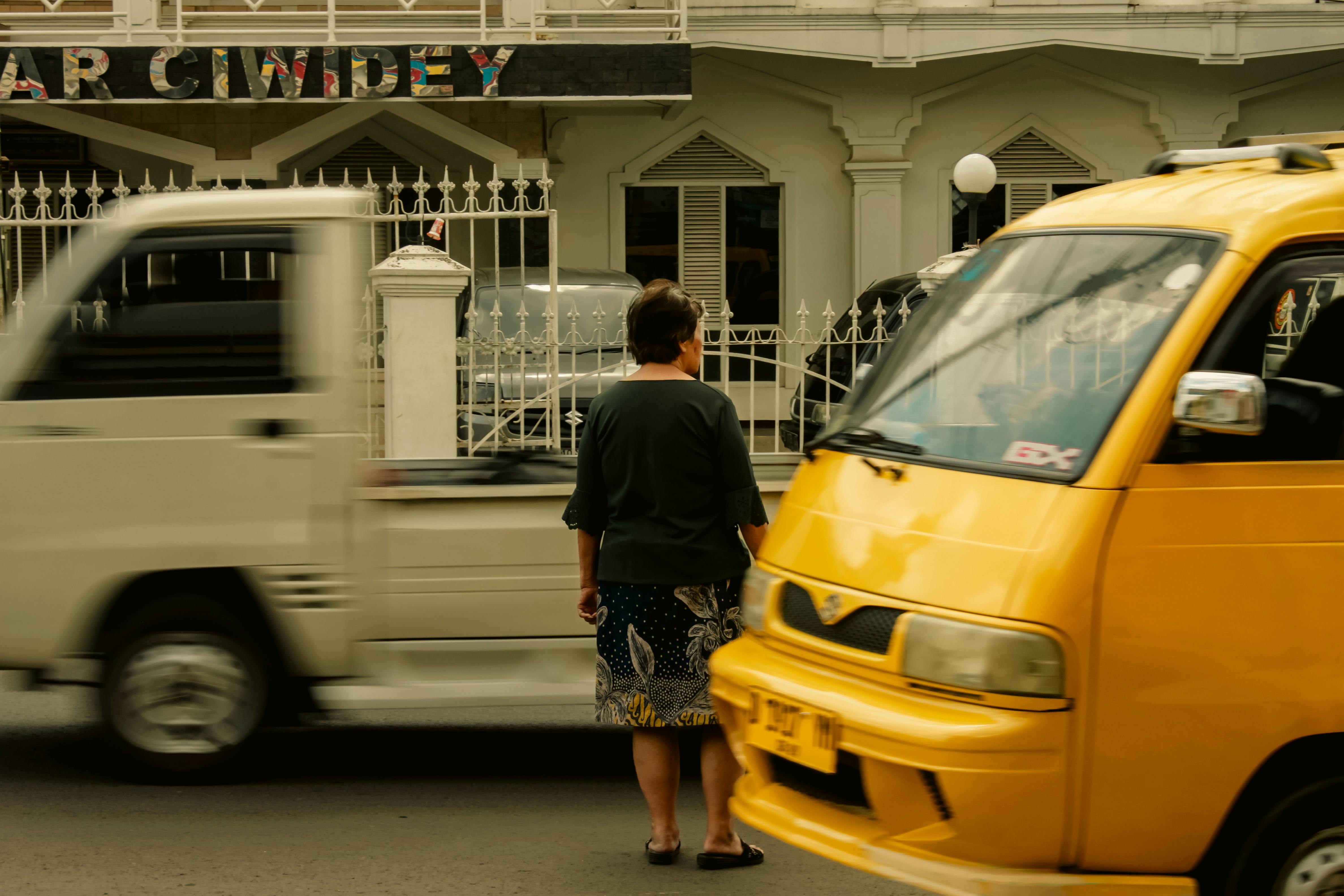 ฟรี หญิงคนหนึ่งยืนอยู่บนถนน ขณะที่รถยนต์สีเหลืองและสีขาวแล่นผ่านไปมาในสภาพแวดล้อมเมือง คลังภาพถ่าย