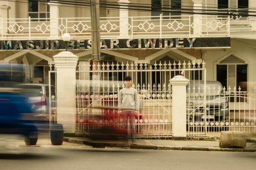 A man stands still in front of the Masjid Besar Ciwidey with blurred traffic passing by, offering a unique urban scene.