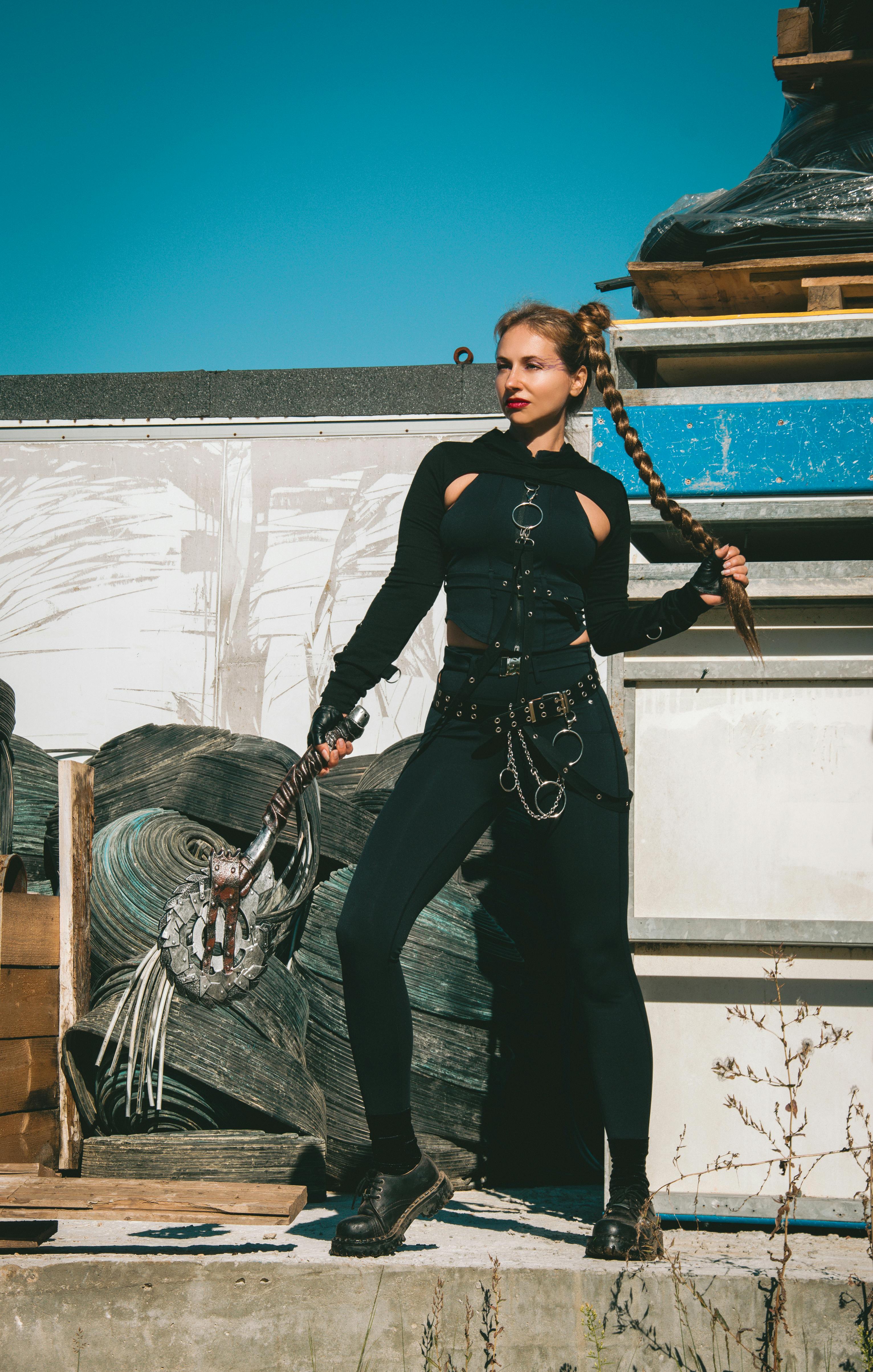 Gratis Una mujer vestida con ropa oscura y tosca adopta una pose fiera empuñando un arma, desprendiendo una atmósfera postapocalíptica. Foto de stock