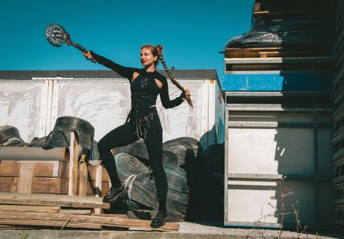 A woman dressed in a warrior costume poses outdoors with braided hair, holding a weapon.