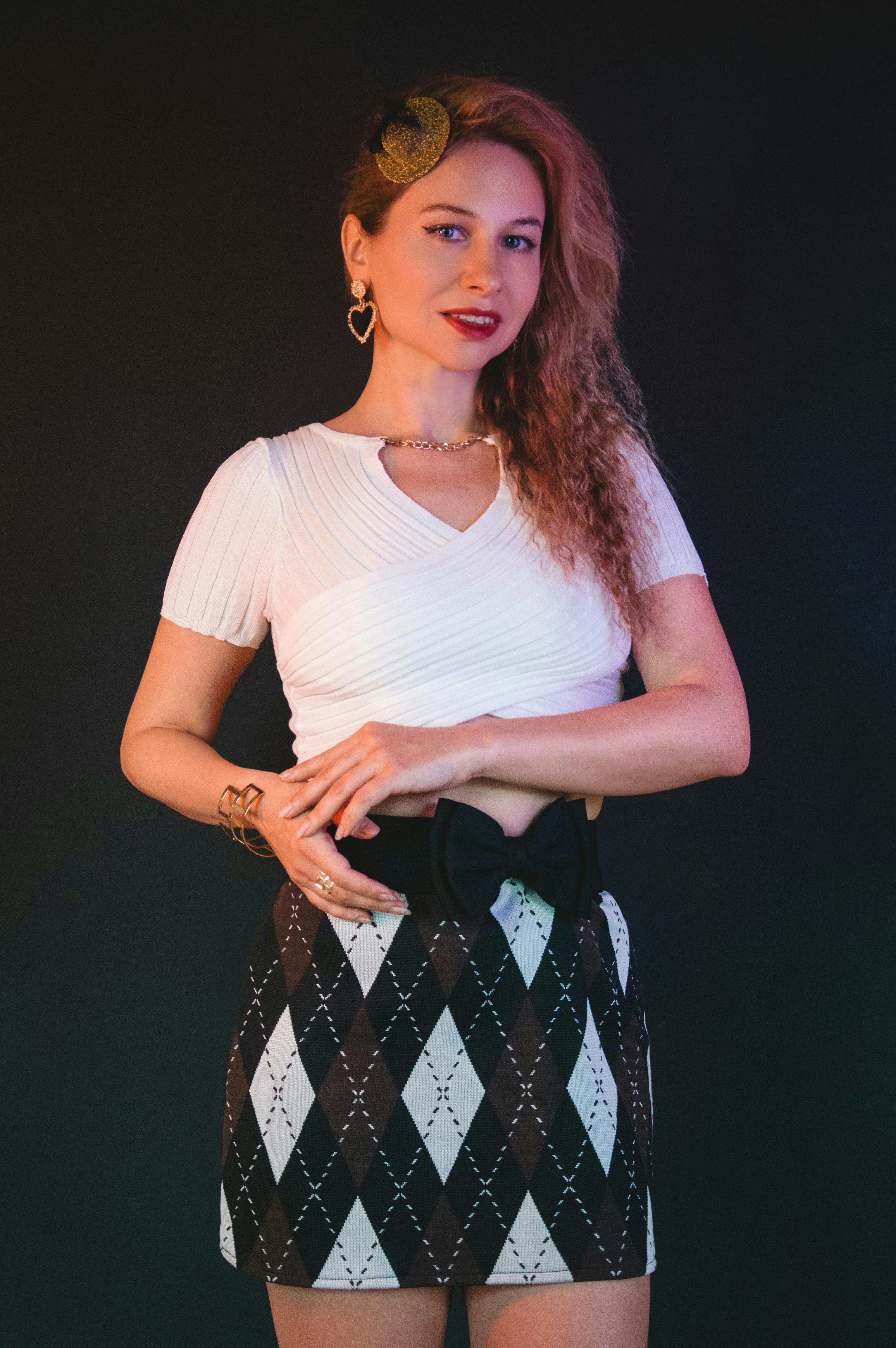 Безкоштовне Стильна жінка у білому топі та спідниці-аргайл впевнено позує на темному тлі. стокове фото