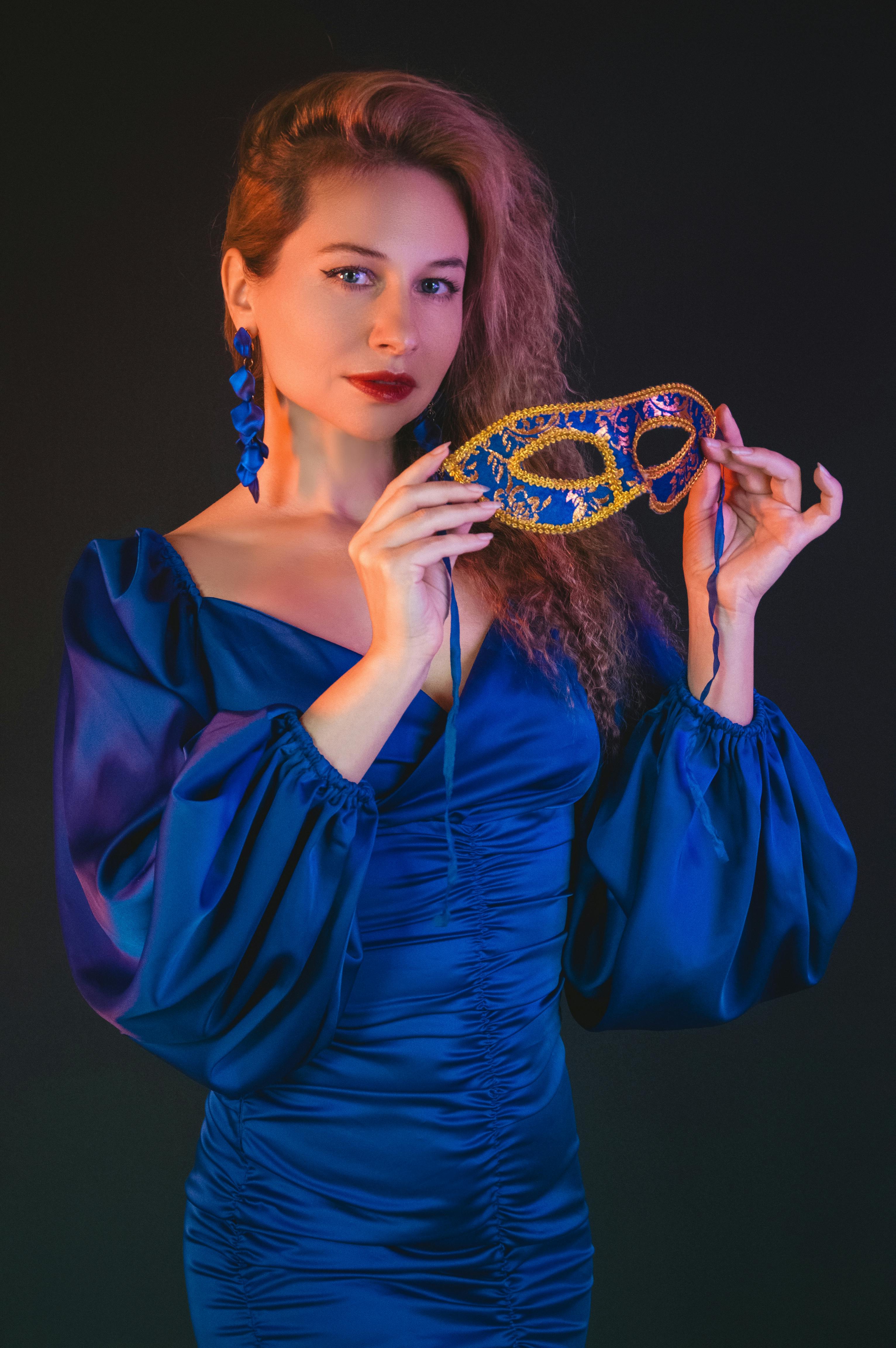 Bezpłatne Kobieta w niebieskiej sukni trzymająca maskę maskaradową na ciemnym tle, emanująca elegancją. Zdjęcie z galerii