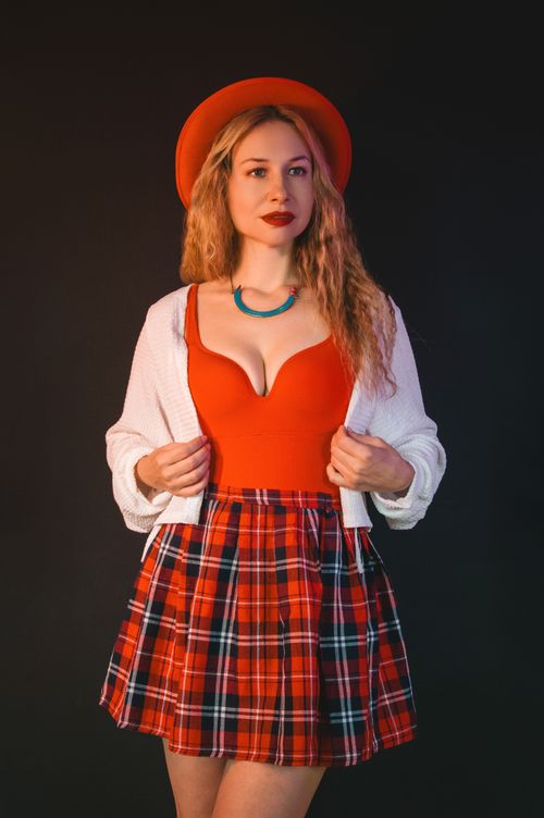 Free Stylish woman in red hat, plaid skirt, and white cardigan posing against black background. Stock Photo