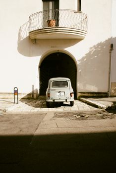 Retro vehicle parked under an archway with a balcony overhead on a sunny street.