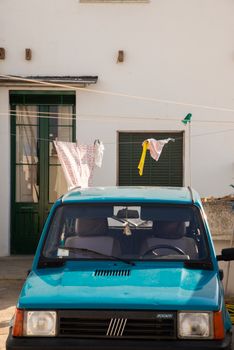 A parked blue car under a clothesline with drying laundry against a white house.