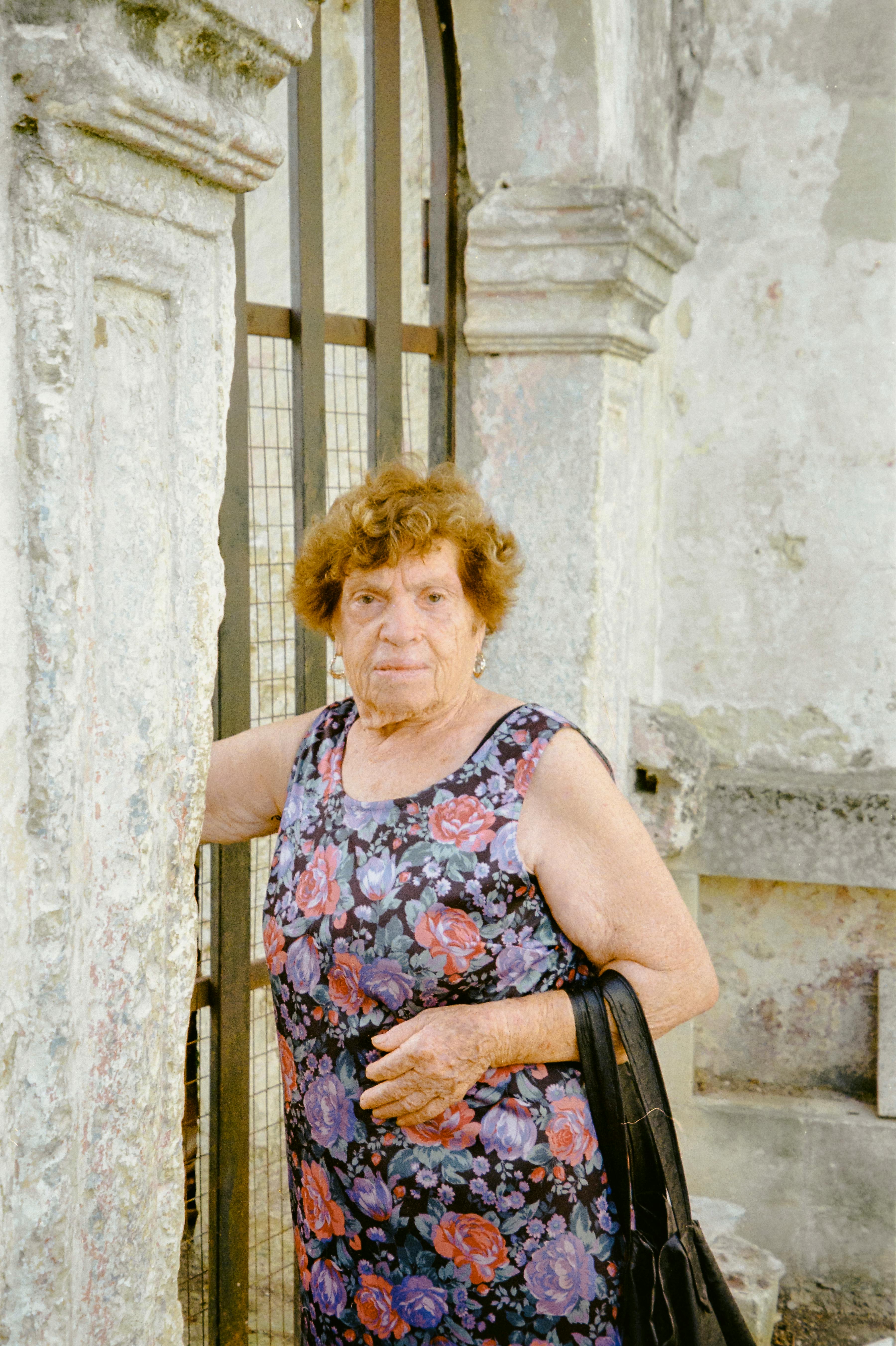 grátis Mulher idosa com vestido florido junto a um arco de pedra rústico, ao ar livre. Foto profissional