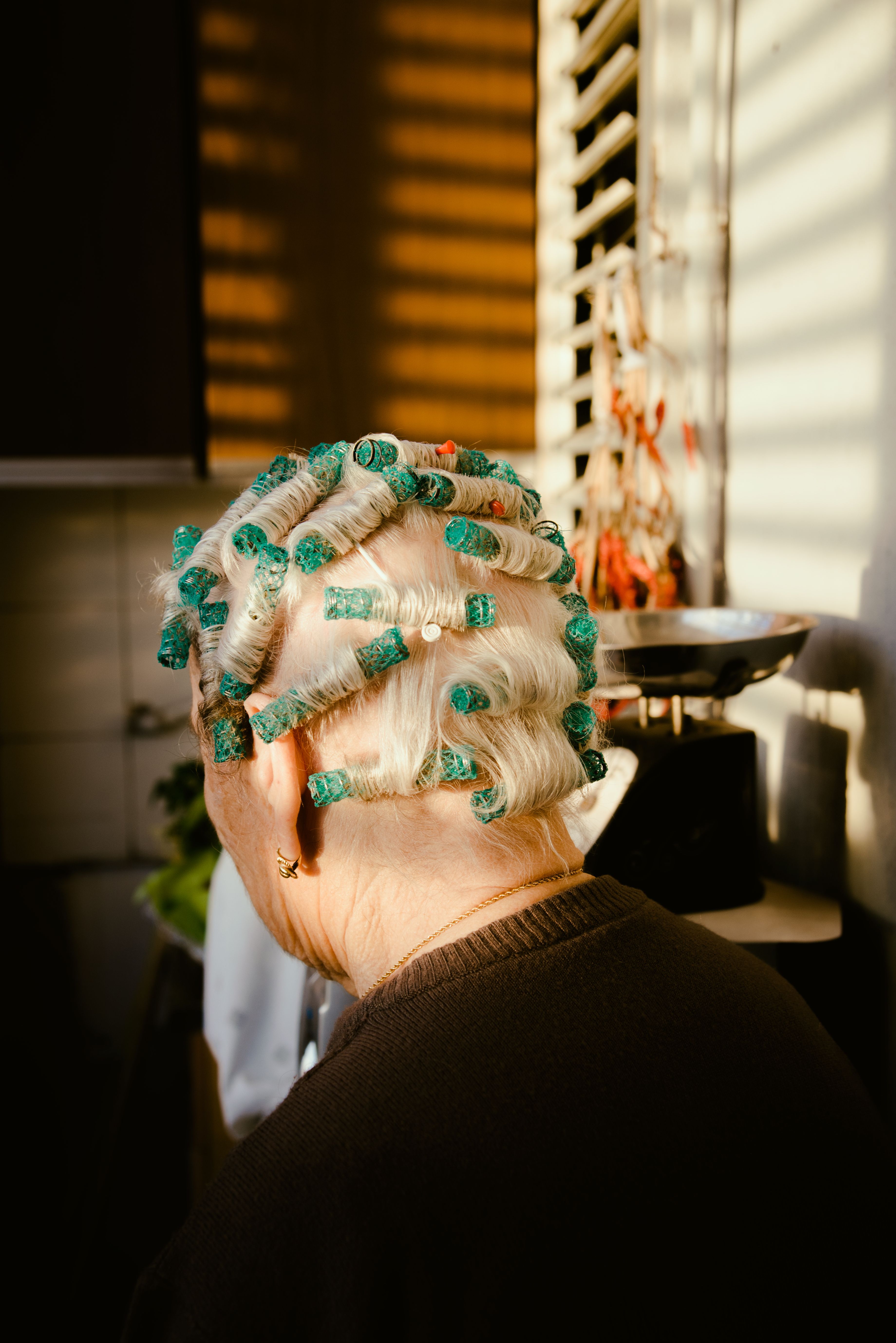 A senior woman with hair curlers enjoys sunlight indoors, evoking warmth and nostalgia.
