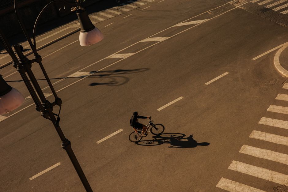 A cyclist rides alone on a sunlit empty street in Lazio, Italy, casting a long shadow.
