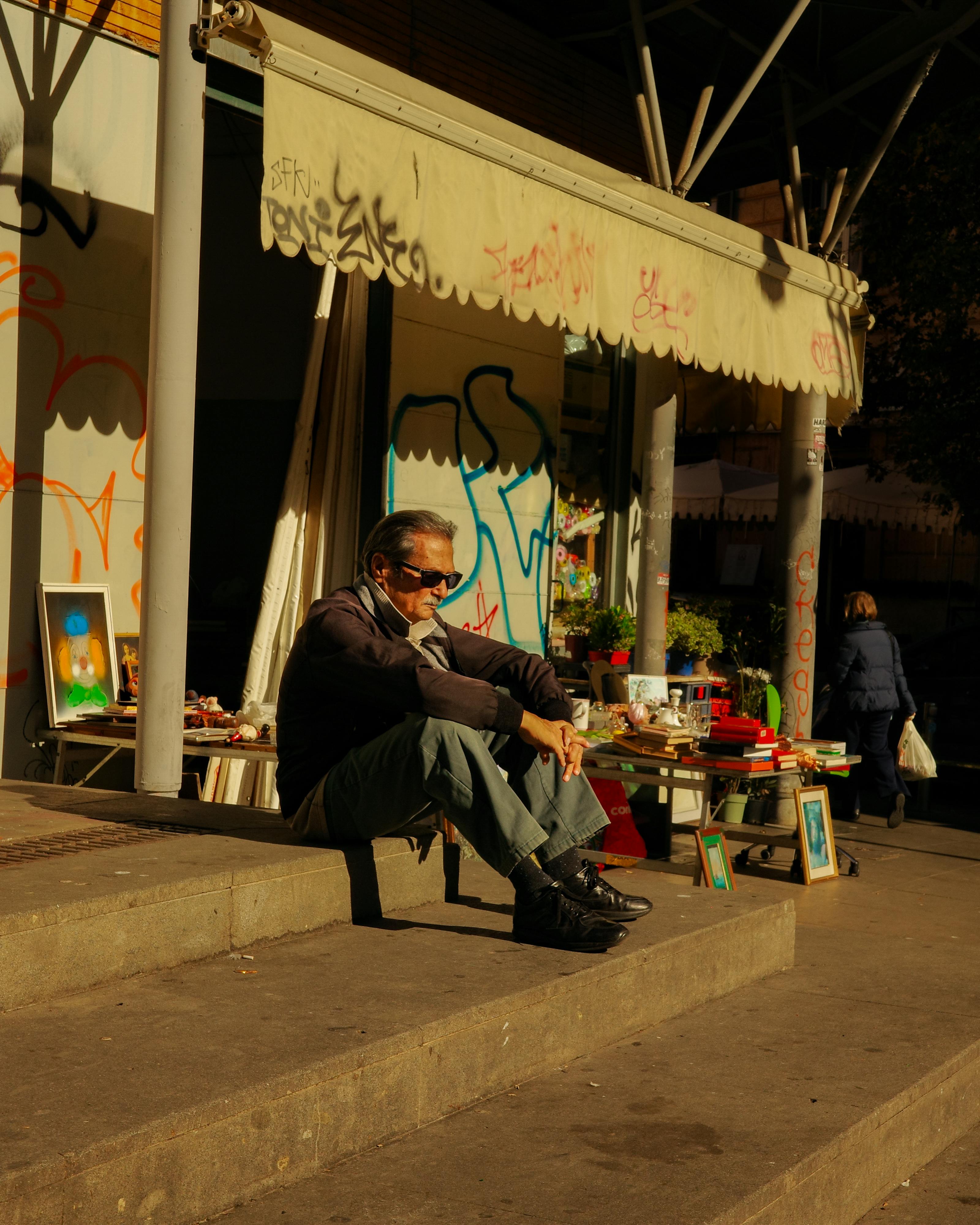 Gratuit Un homme âgé, lunettes de soleil sur le nez, se détend sur les marches d'un marché de rue animé du Latium, sous un soleil radieux. Photos