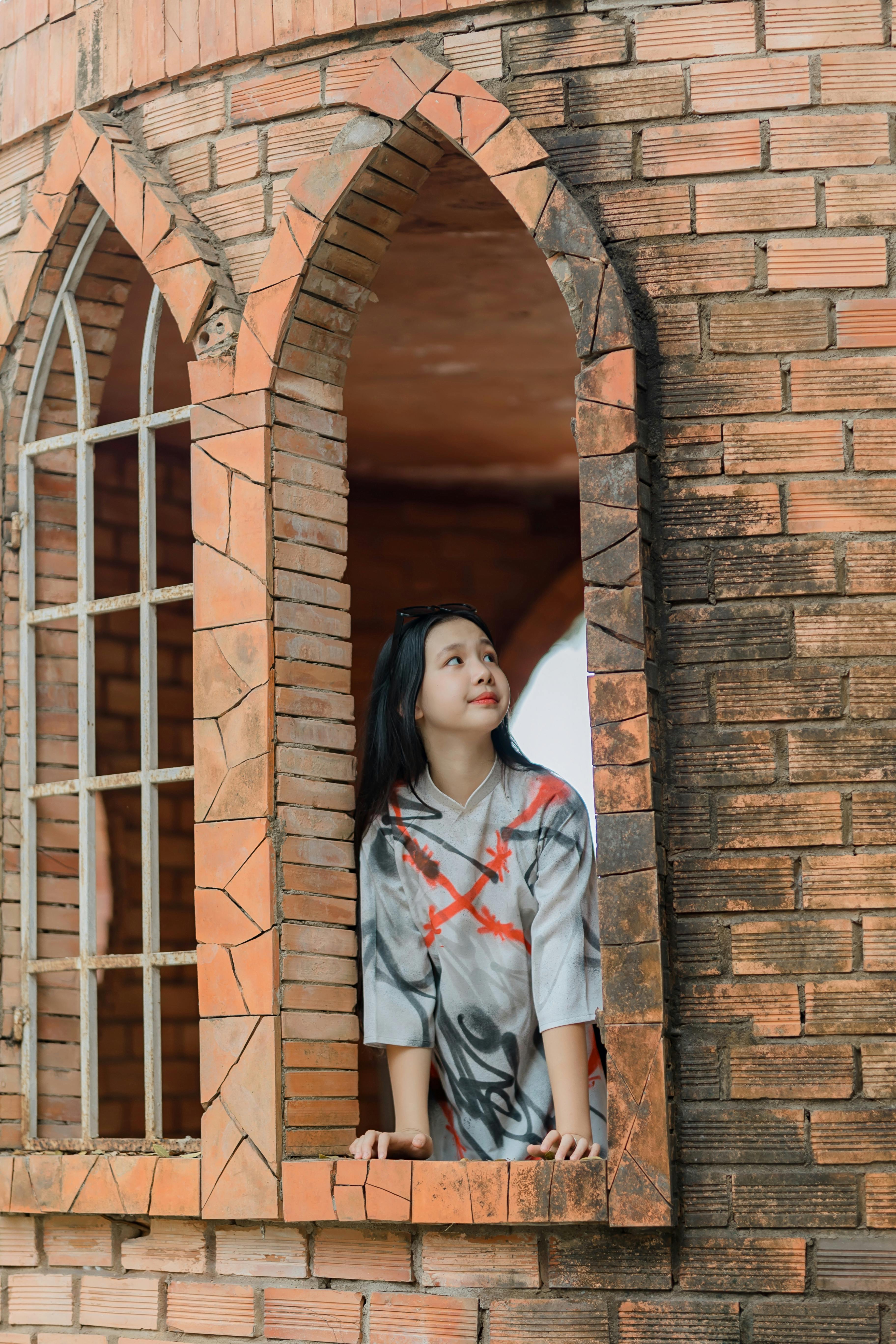 De franc Una dona jove s'acosta a una finestra rústica de maó, mirant pensativa. Foto d'estoc