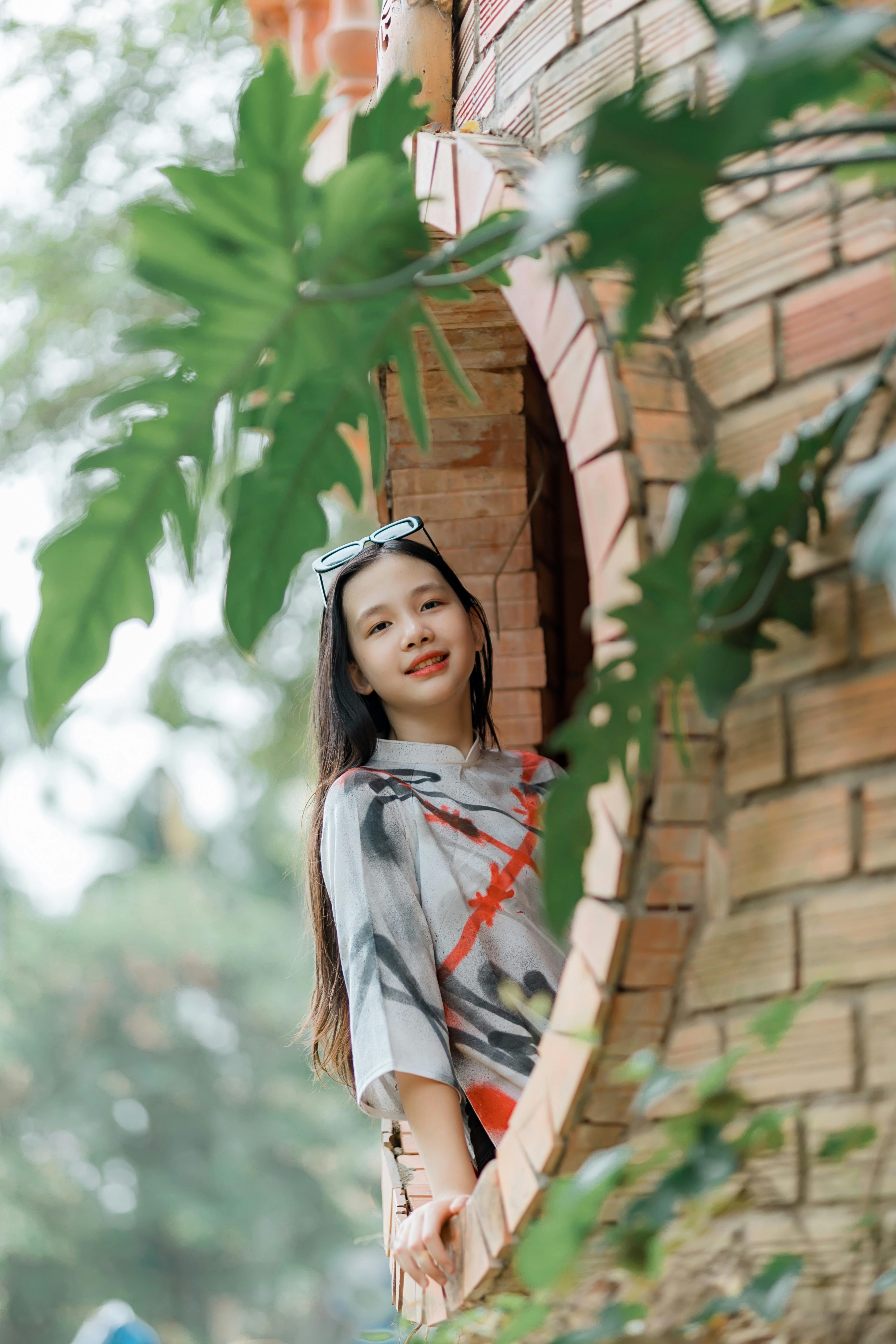 Ücretsiz Yeşillikler içinde, yuvarlak tuğla bir pencerenin önünde poz veren, rengarenk kıyafetler giymiş genç bir kadın. Stok Fotoğraflar