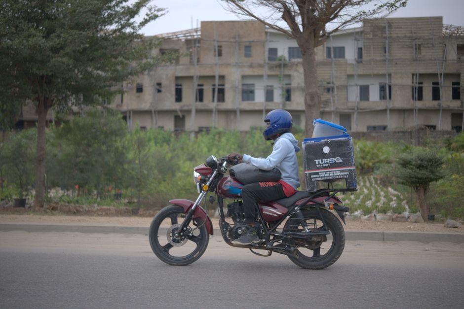 Elderly Black Nigerian woman in a colourful adire blouse opening her front door in a Nigerian neighbourhood to receive a box of groceries from a delivery person, warm afternoon light, photorealistic