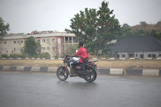 A man riding a motorcycle on a city street in Abuja, Nigeria with buildings and trees in the background.