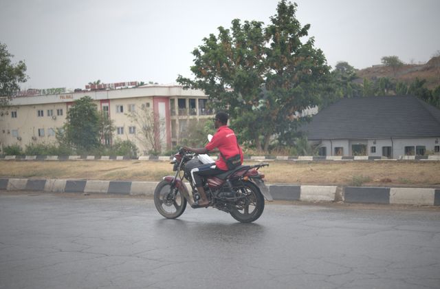 A man riding a motorcycle on a city street in Abuja, Nigeria with buildings and trees in the background. — arriving in Abuja