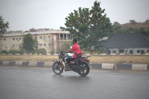 Free A man riding a motorcycle on a city street in Abuja, Nigeria with buildings and trees in the background. Stock Photo