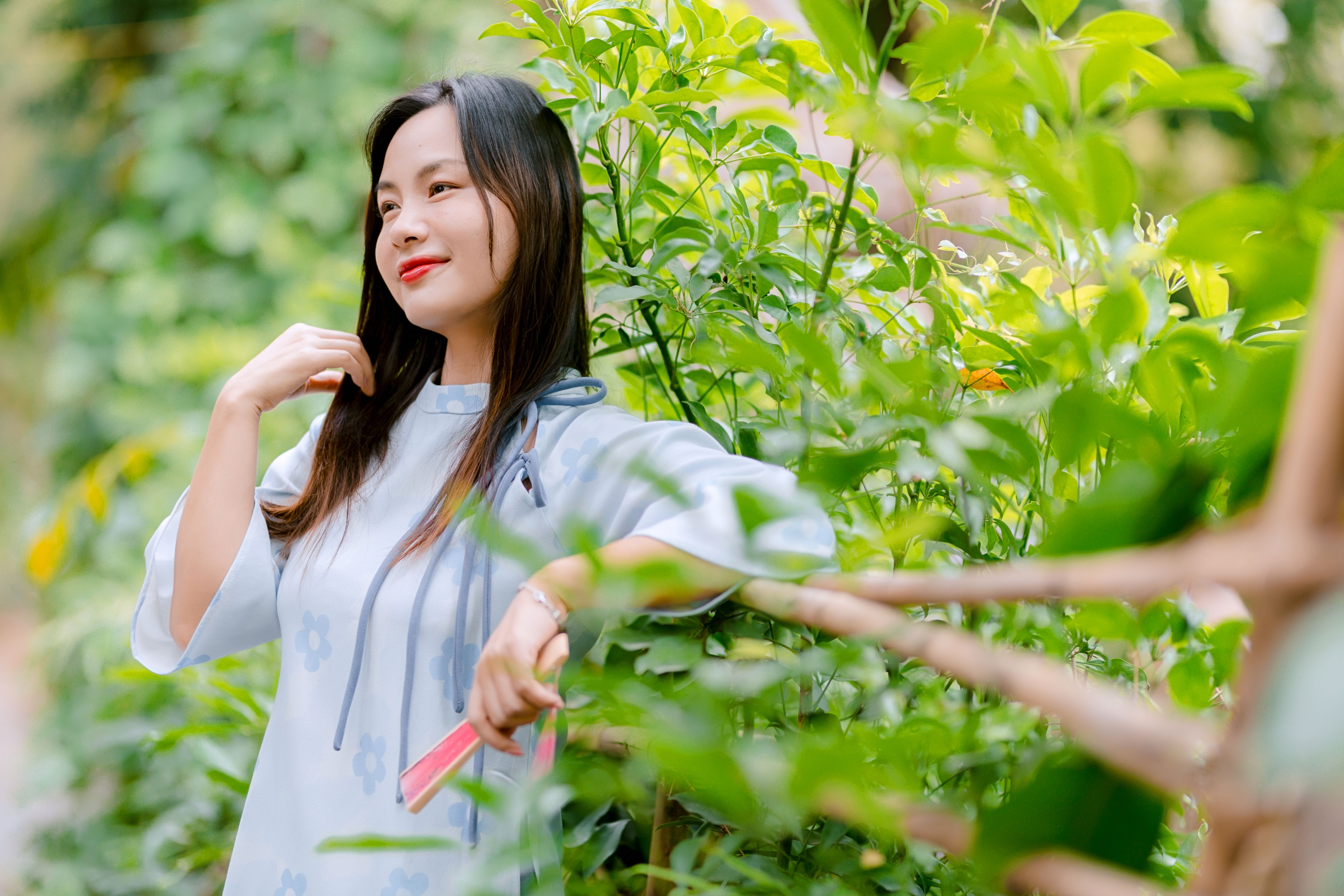 A serene portrait of a woman in a light blue dress standing among vibrant green leaves outdoors.