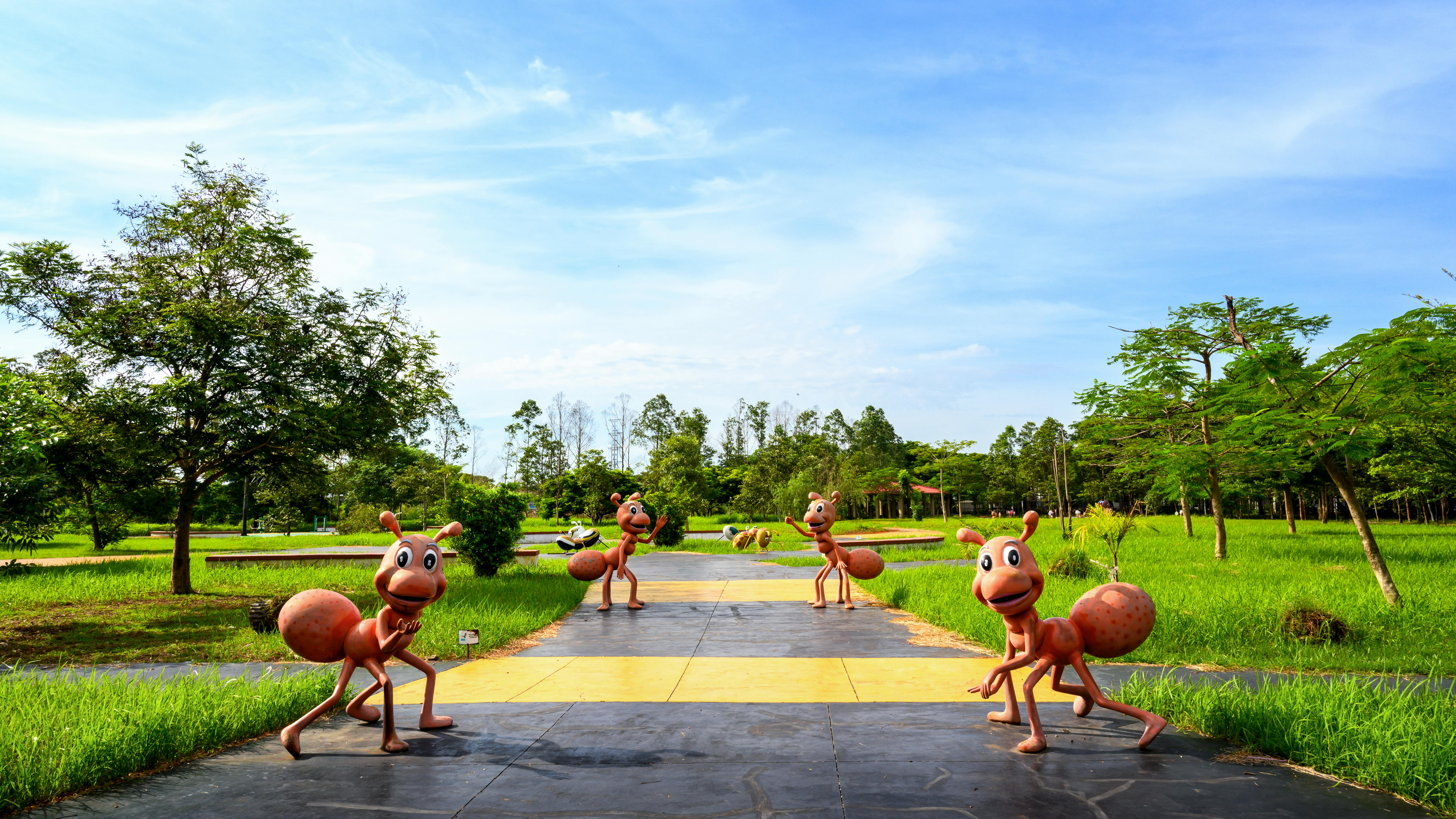 Colorful ant sculptures line a sunny park pathway amidst lush greenery.