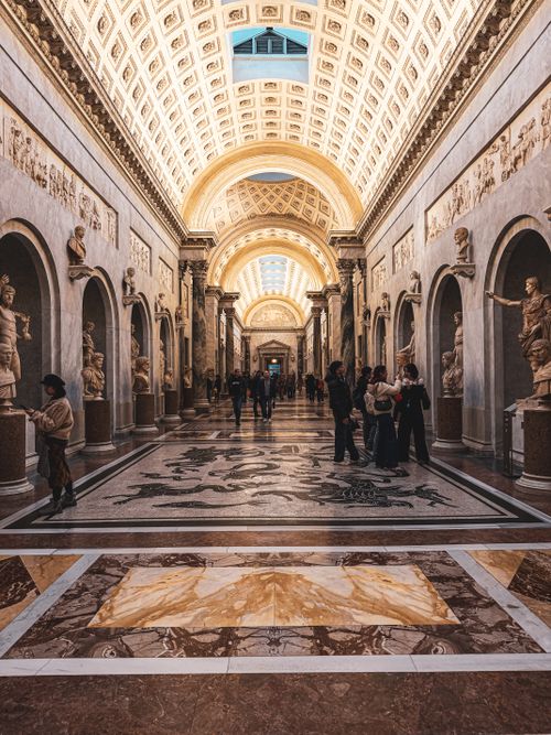 Free View of a grand museum hallway with classical sculptures and intricate ceilings, filled with visitors. Stock Photo