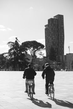 Two men cycling in a city square with modern architecture in black and white.