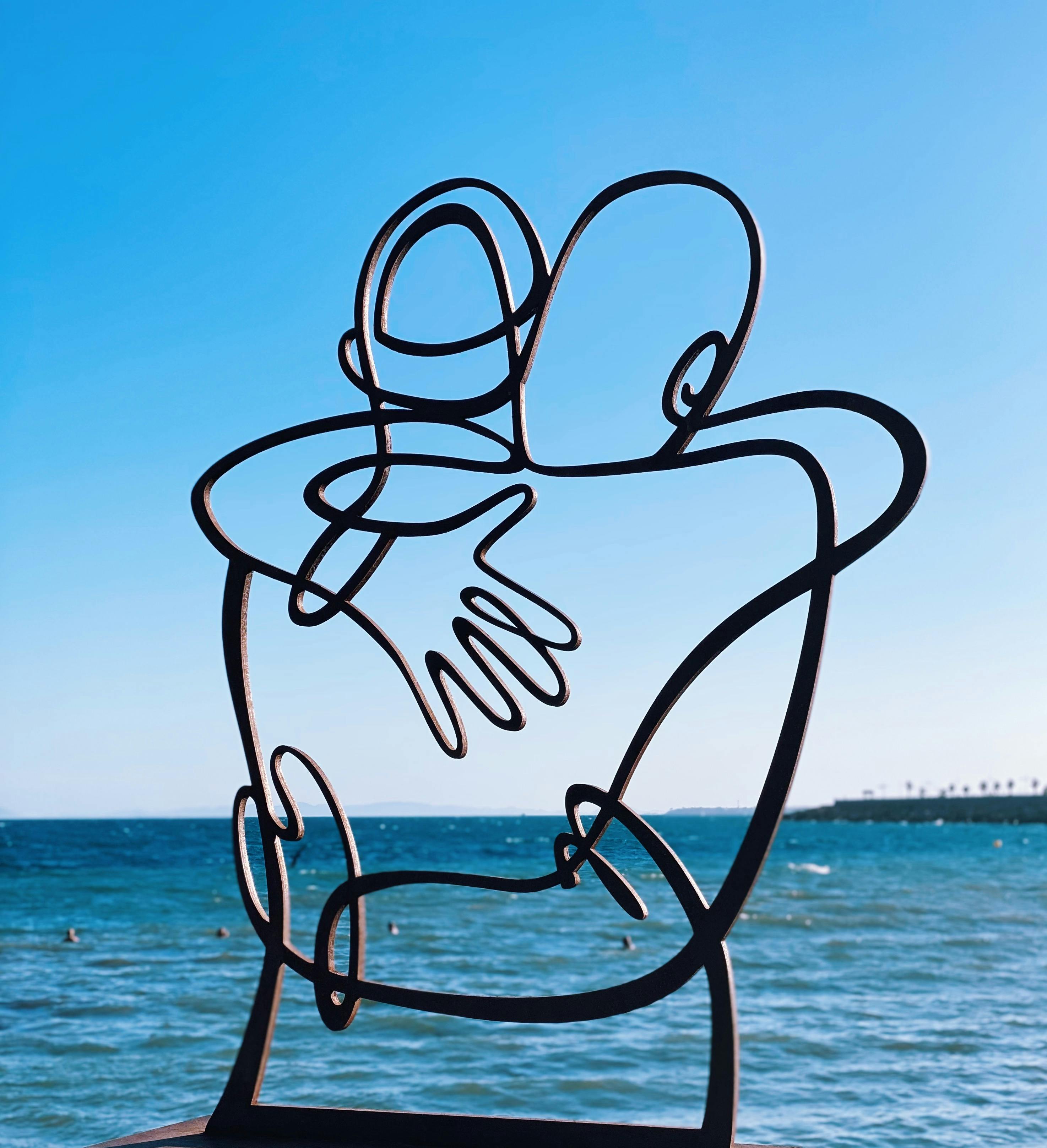 Gratuit Une sculpture abstraite en métal représentant des silhouettes enlacées se détachant sur un fond de mer et de ciel d'un bleu limpide. Photos
