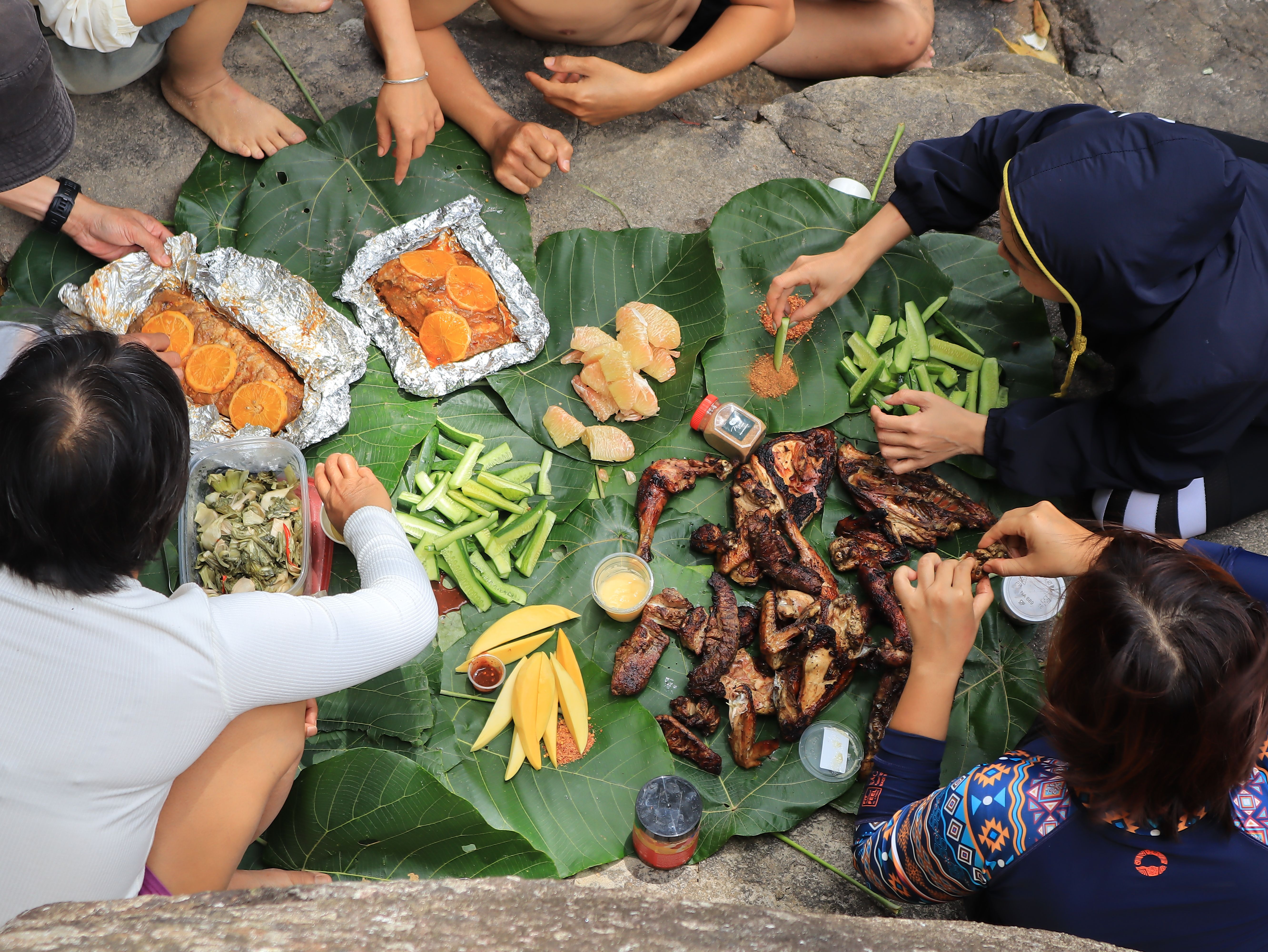 Gathering of people enjoying grilled food and fruits on banana leaves outdoors.