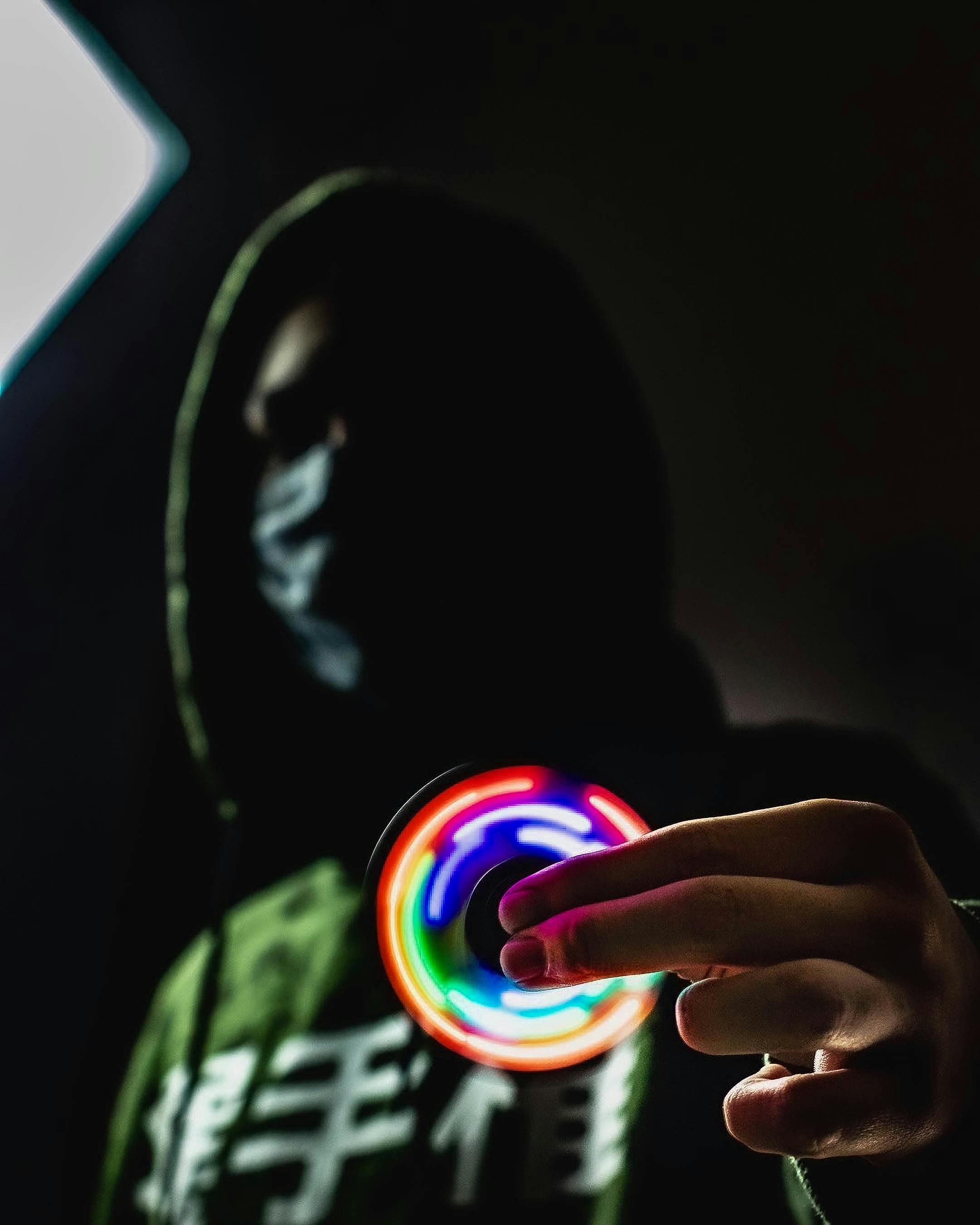 Gratis Una misteriosa figura mascherata, in penombra, regge in mano uno spinner a LED dai colori vivaci. Foto a disposizione