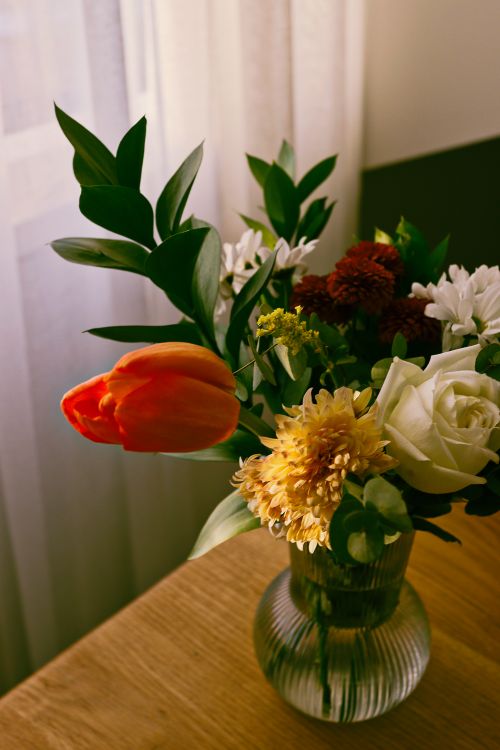Free A vibrant floral arrangement featuring tulip, rose, and chrysanthemum in a glass vase on a wooden table. Stock Photo