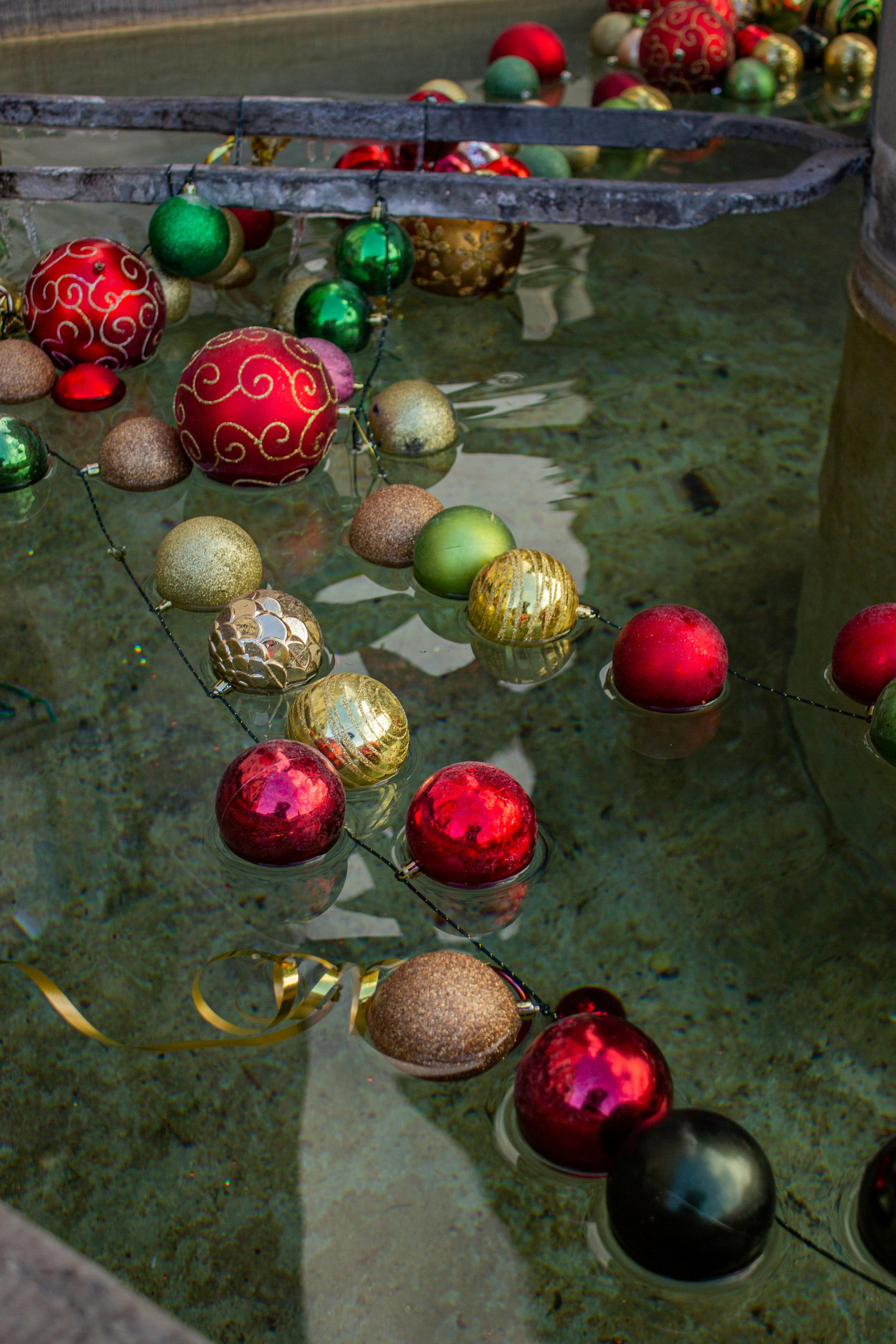 Festive red, gold, and green Christmas balls float in a clear water fountain, depicting holiday cheer.