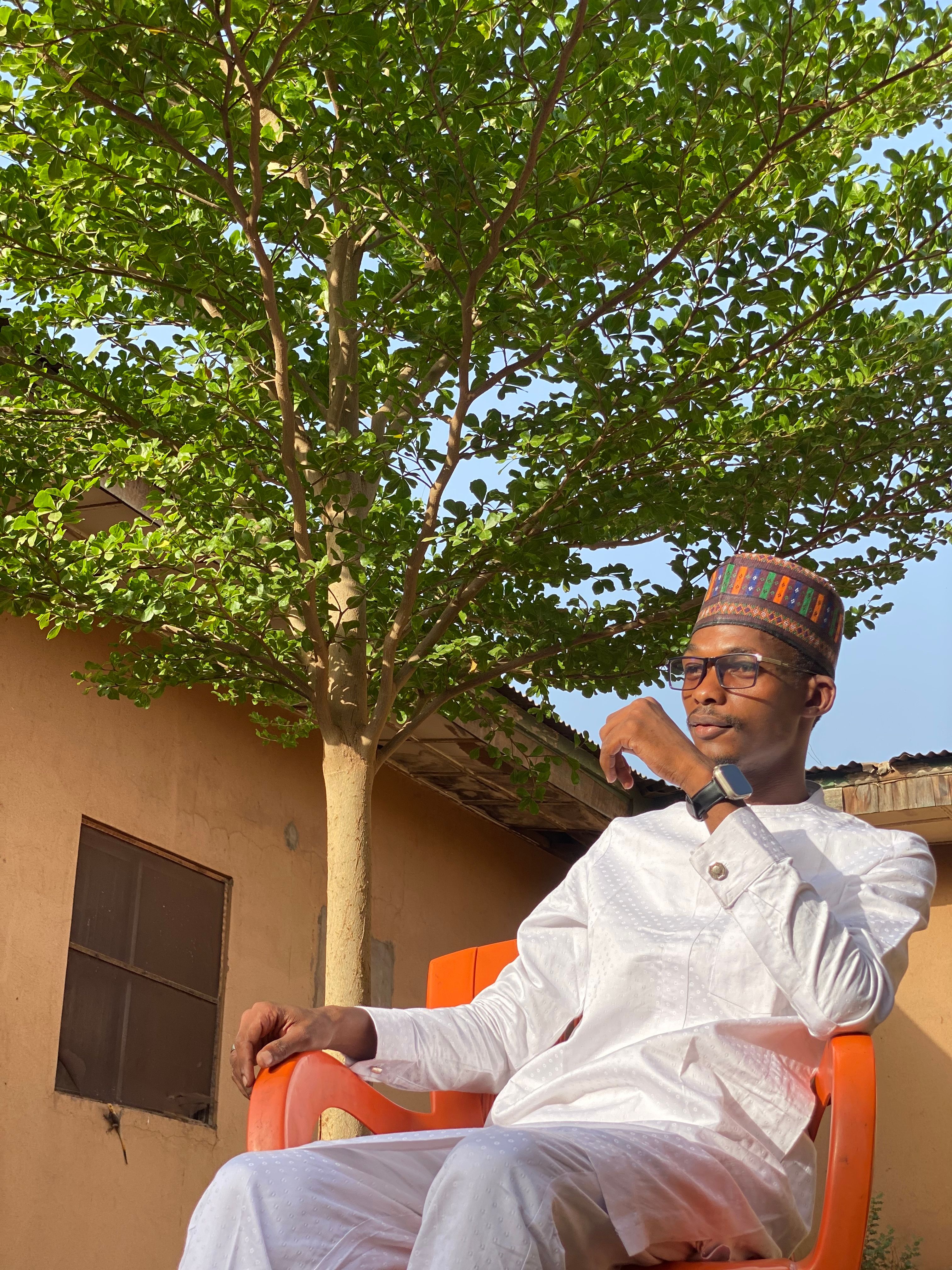 A young man wearing traditional Nigerian attire sits outdoors under a tree.