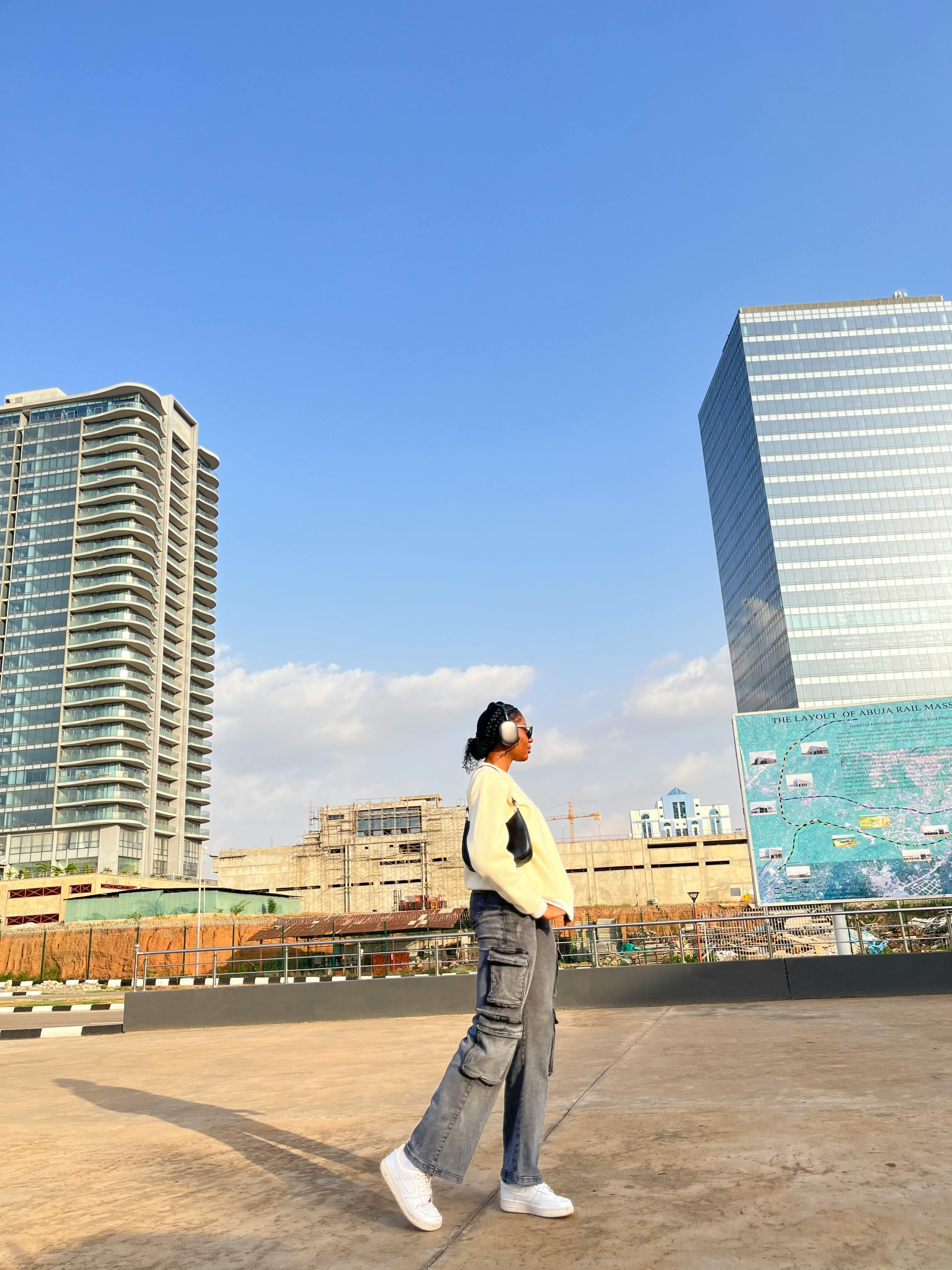 Kostenlos Ein junger Erwachsener mit Kopfhörern steht vor Wolkenkratzern vor einem strahlend blauen Himmel in Abuja, Nigeria. Stock-Foto