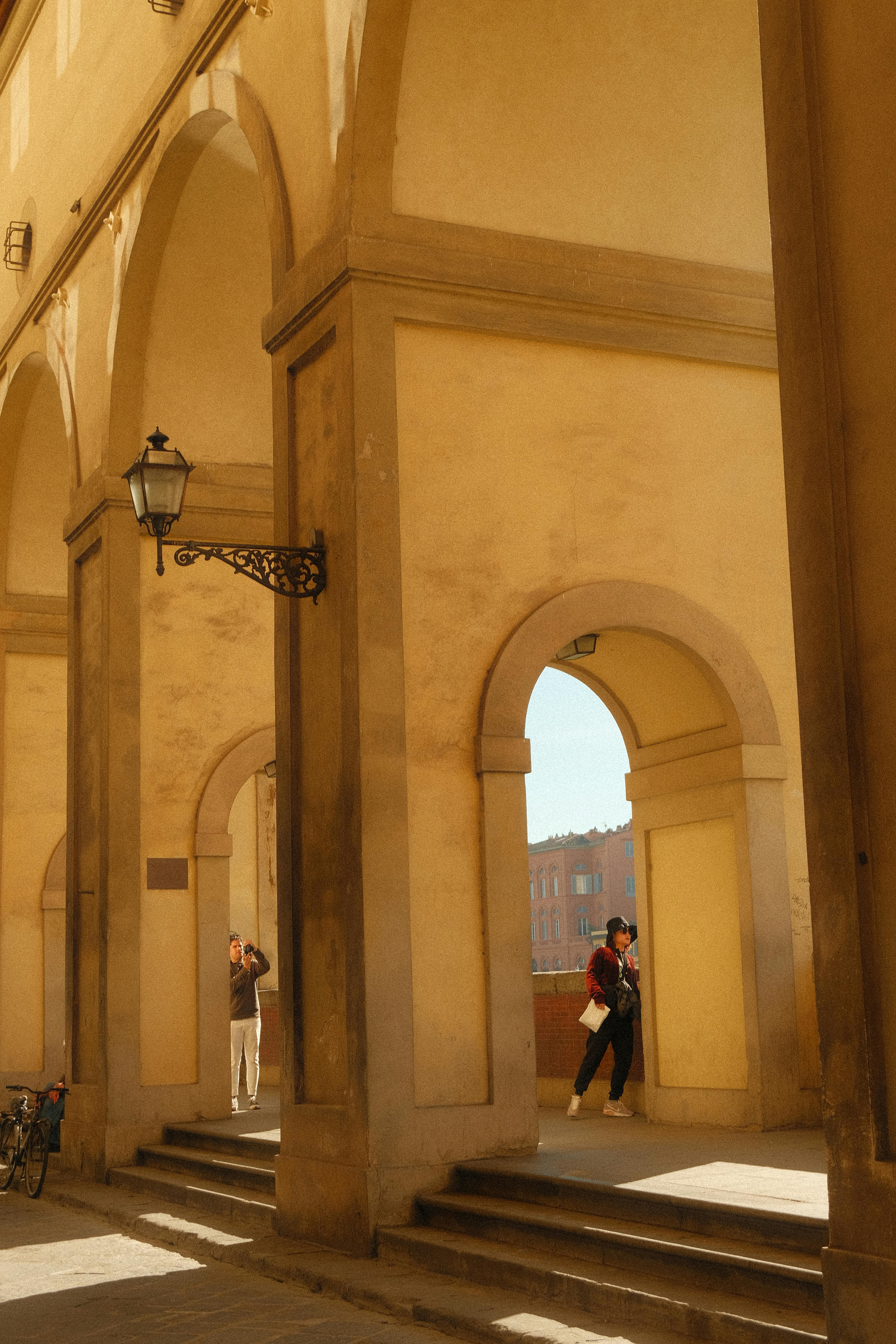 Gratuit Une scène architecturale avec des personnes déambulant sous des arcades historiques dans un environnement urbain baigné de soleil. Photos