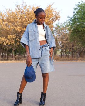Stylish woman in denim jacket and shorts holding a cap outdoors.