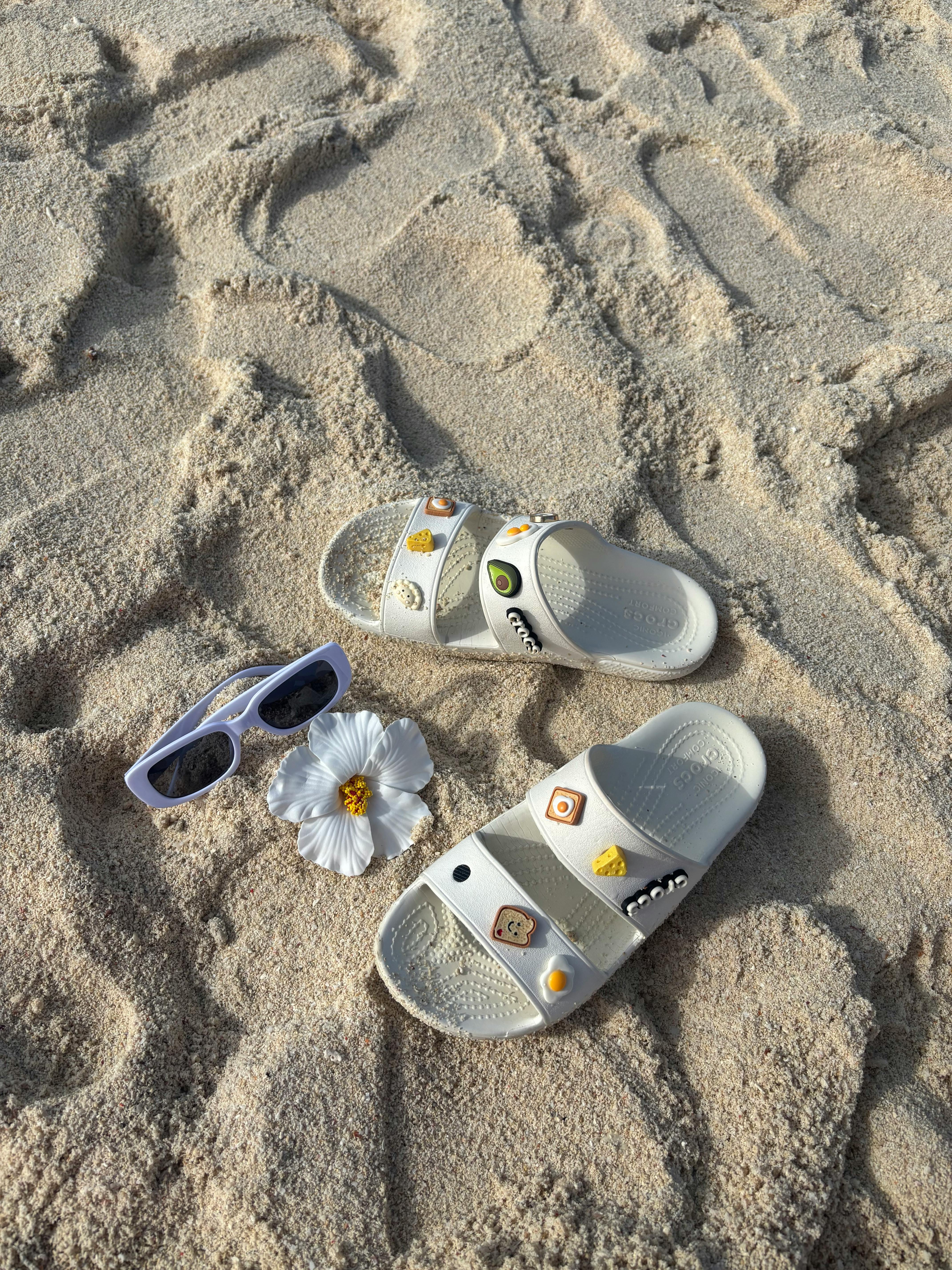 Ücretsiz Yaz havasını yansıtan, kumlu bir plajda, beyaz sandaletler, üzerlerinde süslemeler, bir çiçek ve güneş gözlüğü. Stok Fotoğraflar