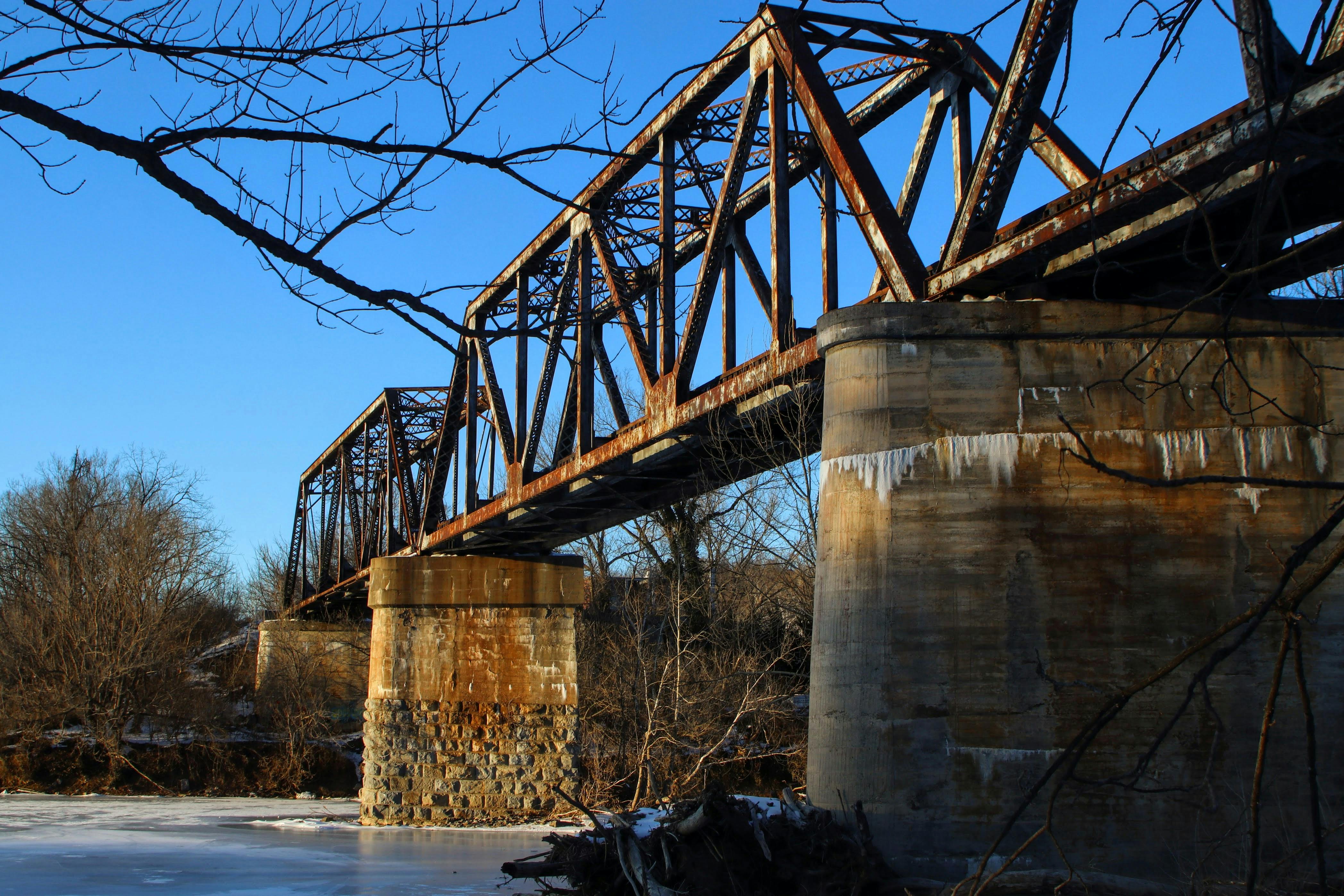 A rusty iron bridge spans over a frozen river on a clear winter day, capturing a serene outdoor scene.