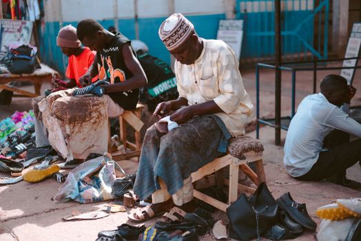 Three shoemakers crafting leather shoes in a vibrant outdoor market in Jos, Nigeria.