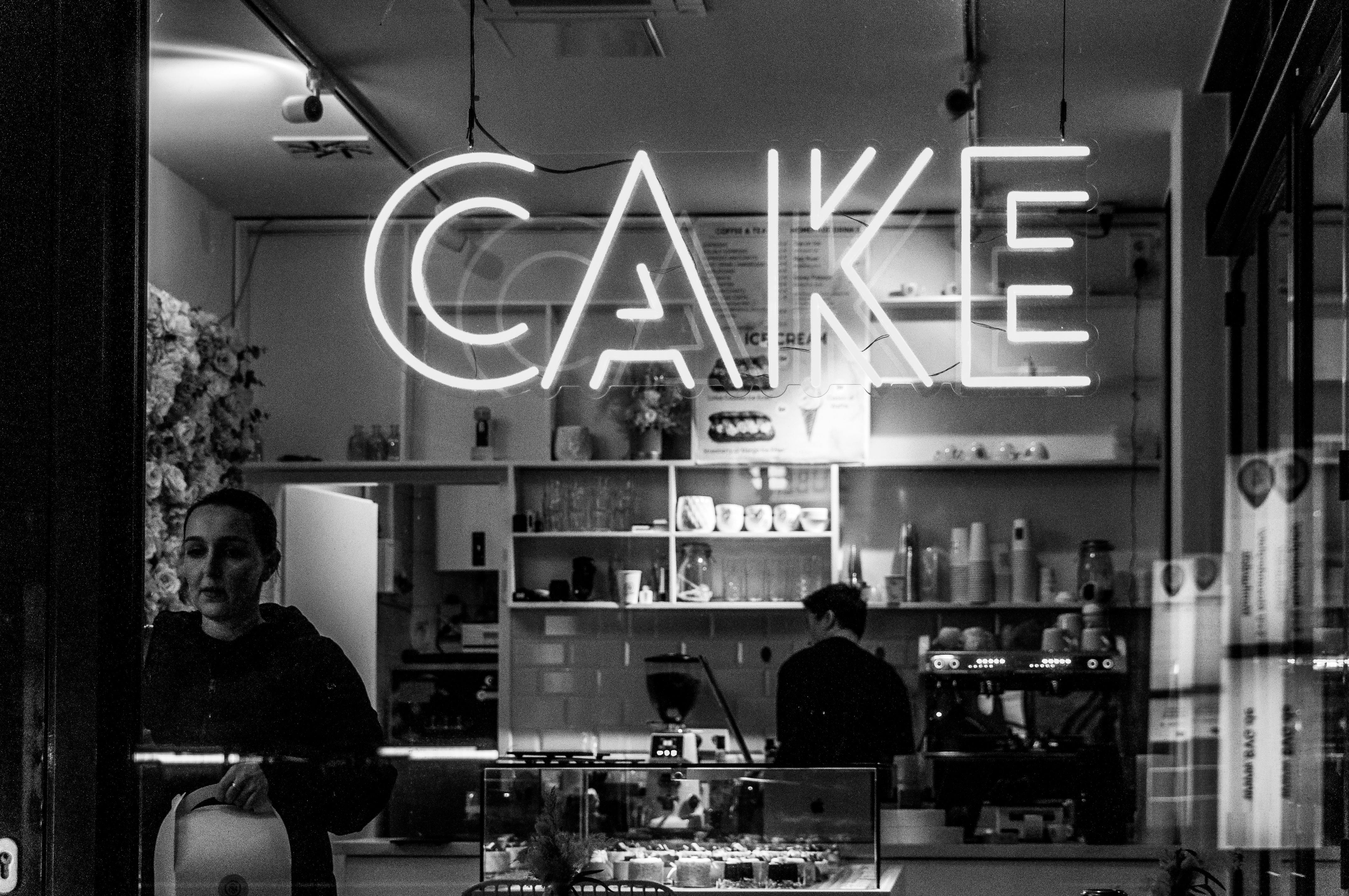 Δωρεάν Μια ασπρόμαυρη φωτογραφία του εσωτερικού ενός ζαχαροπλαστείου με μια νέον πινακίδα που γράφει «CAKE». Φωτογραφία από στοκ φωτογραφιών