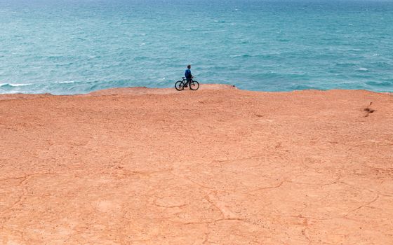 A lone cyclist rides along an arid cliff edge with a vast ocean view in Natal, Brazil.