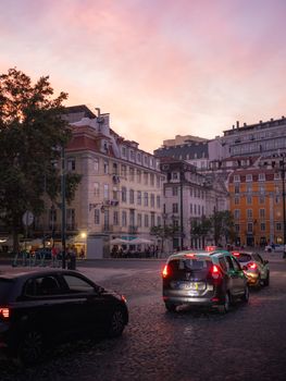 Evening traffic amidst Lisbon's historic architecture under a vibrant pink sky, capturing urban life.