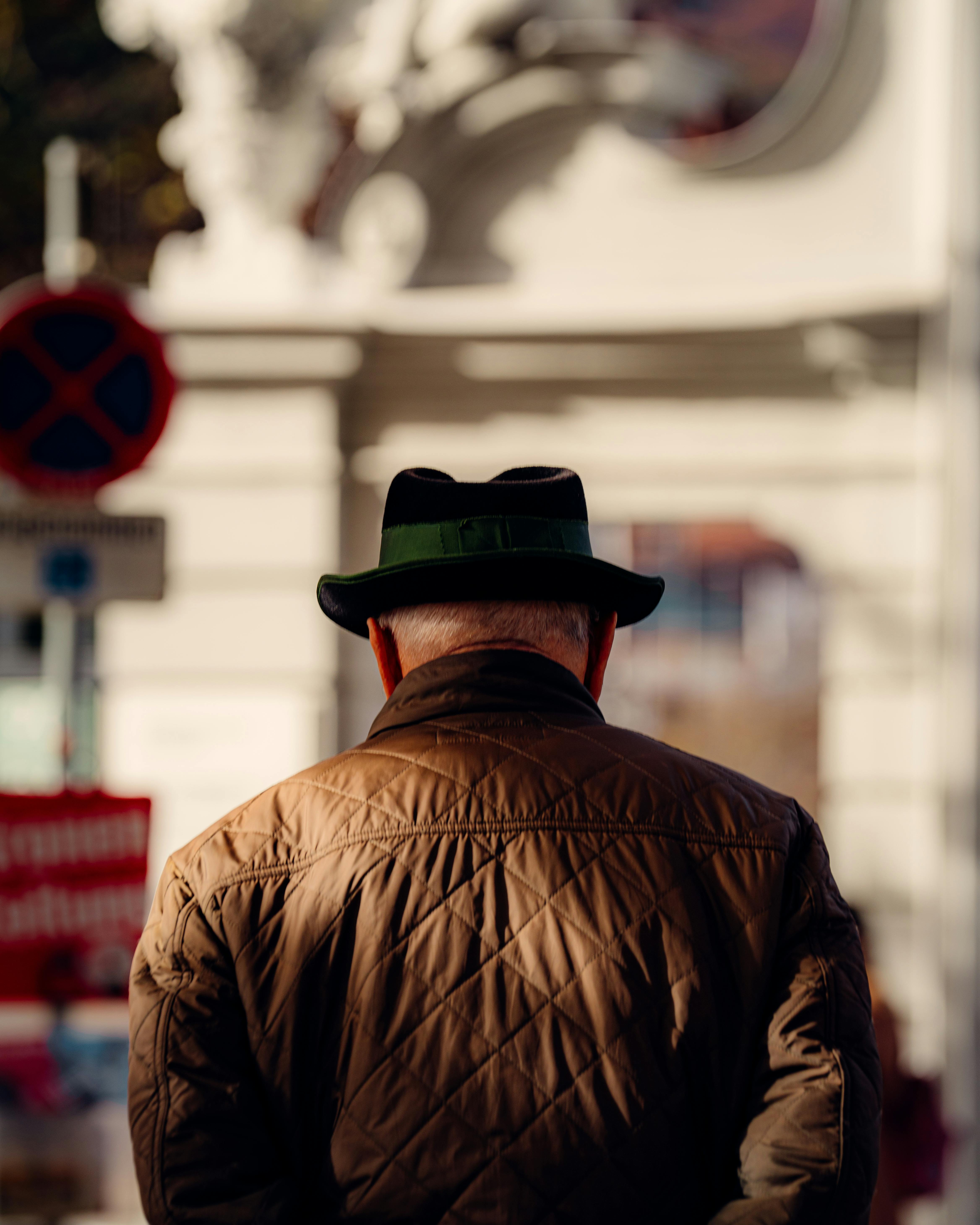 Gratis Un uomo anziano con giacca e cappello marroni cammina in un ambiente urbano illuminato dal sole durante l'autunno. Foto a disposizione