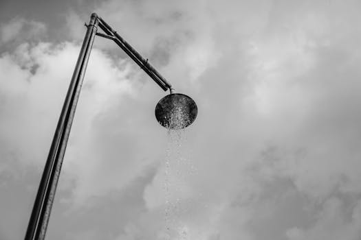 A black and white depiction of an outdoor shower with water against a cloud-filled sky.