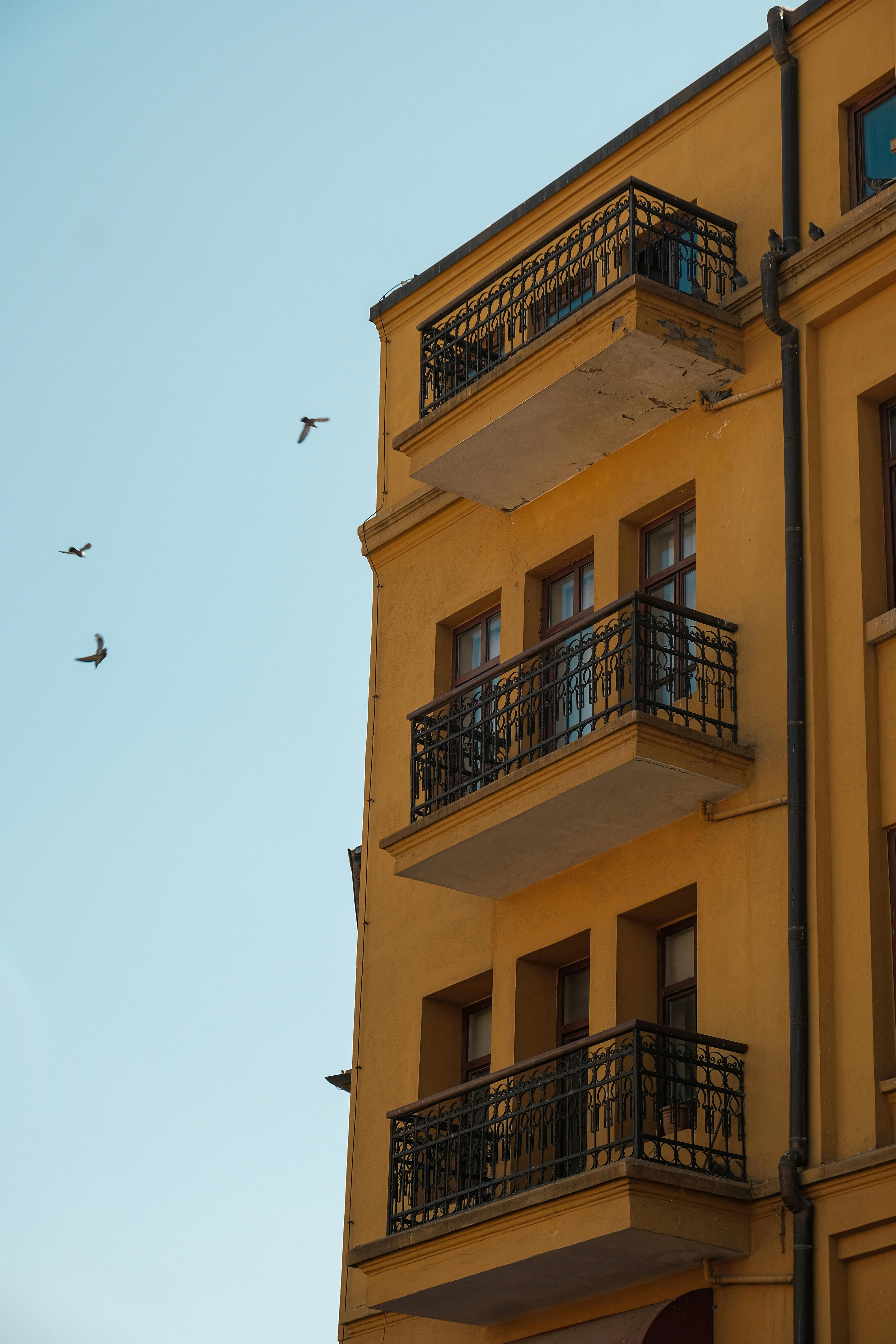 grátis Um belo edifício amarelo com varandas contra um céu claro em Ancara, Turquia, com pássaros voando ao fundo. Foto profissional