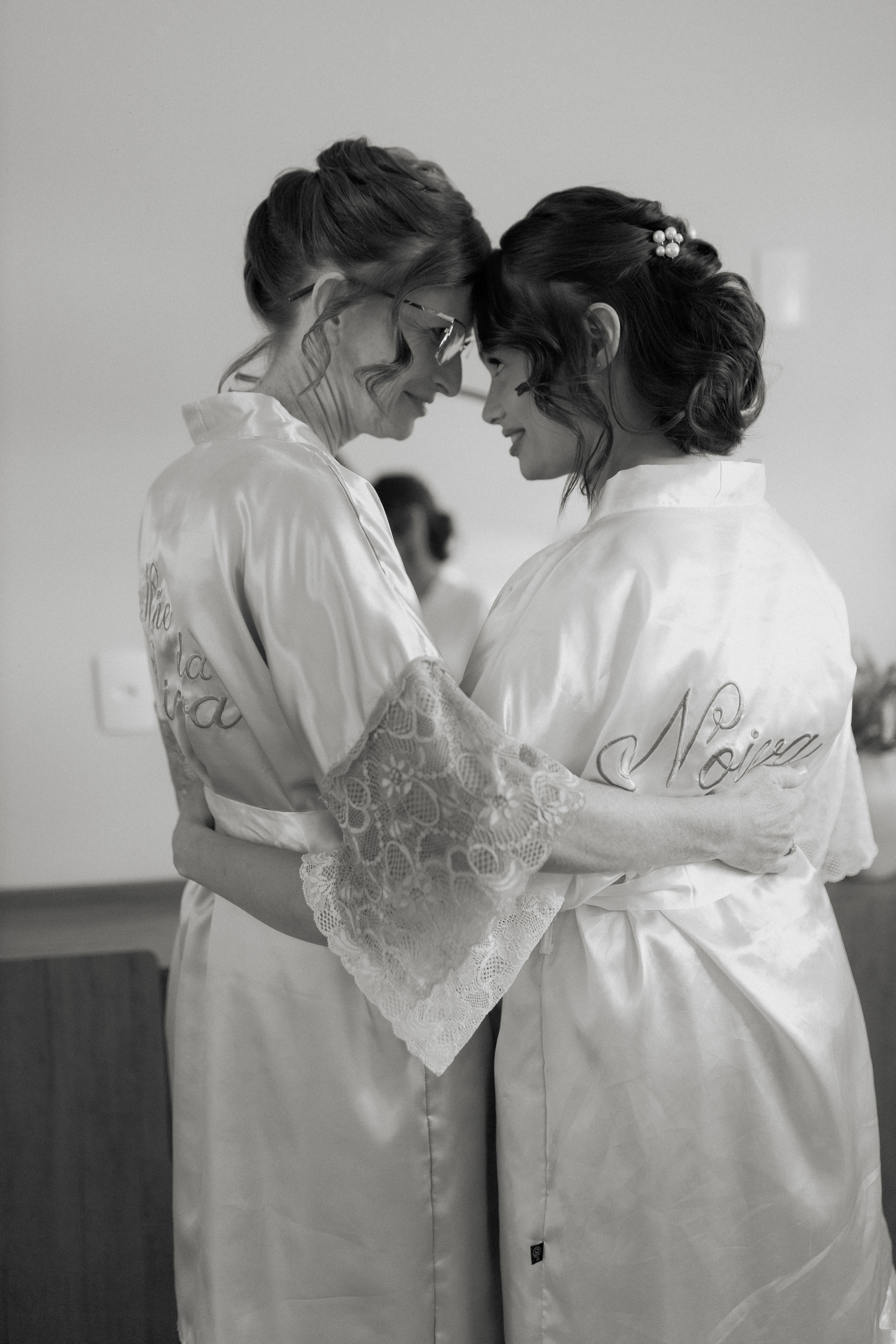 Two women in bridal robes share an intimate embrace before a wedding, conveying warmth and friendship.