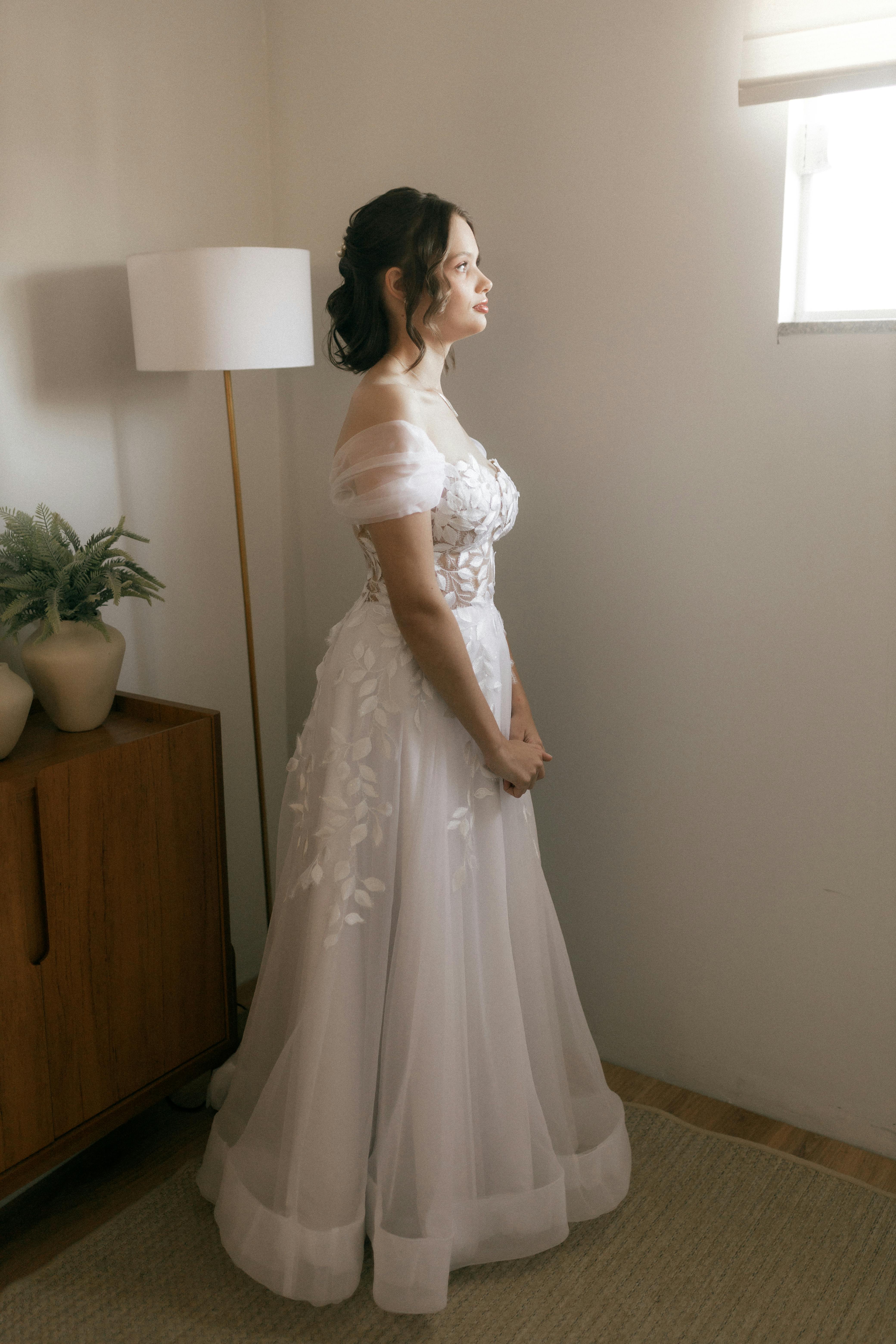 Gratis La sposa, in un elegante abito bianco, guarda fuori dalla finestra di una stanza illuminata da una luce soffusa, catturando un'atmosfera serena e contemplativa. Foto a disposizione