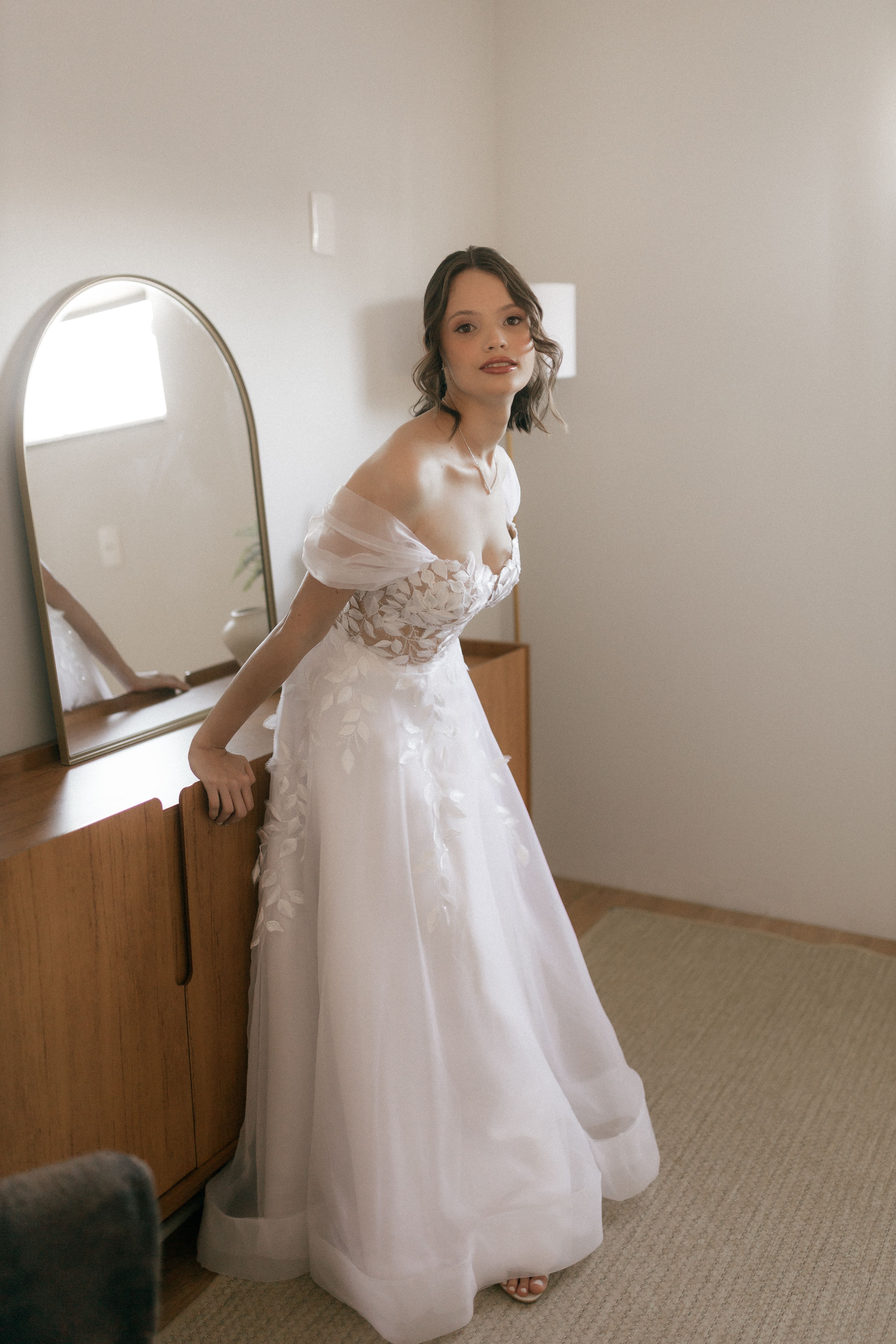 Bride posing gracefully indoors, wearing a delicate white gown in a softly lit room.