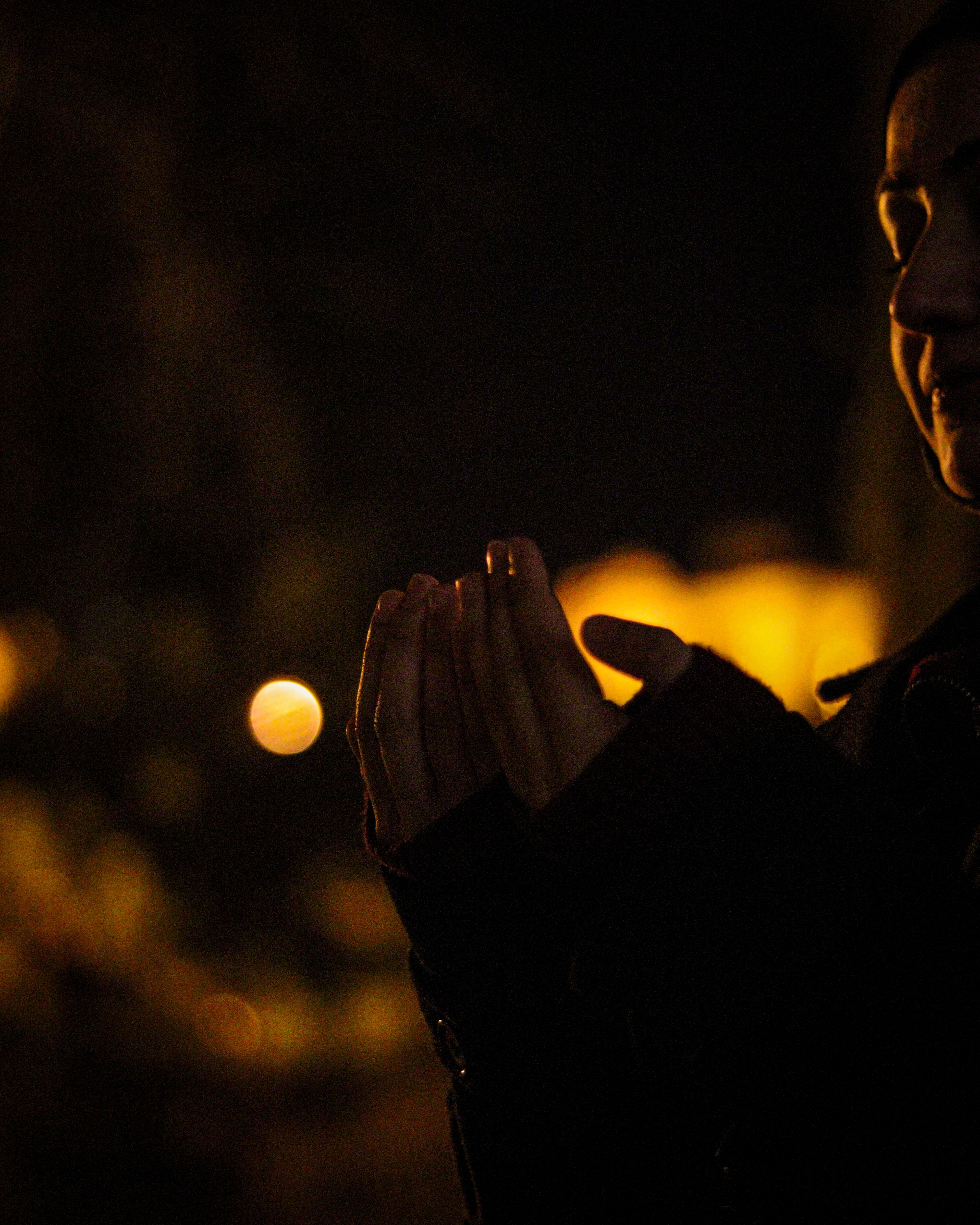 Silhouette of person with hands clasped in prayer amidst night bokeh lights.